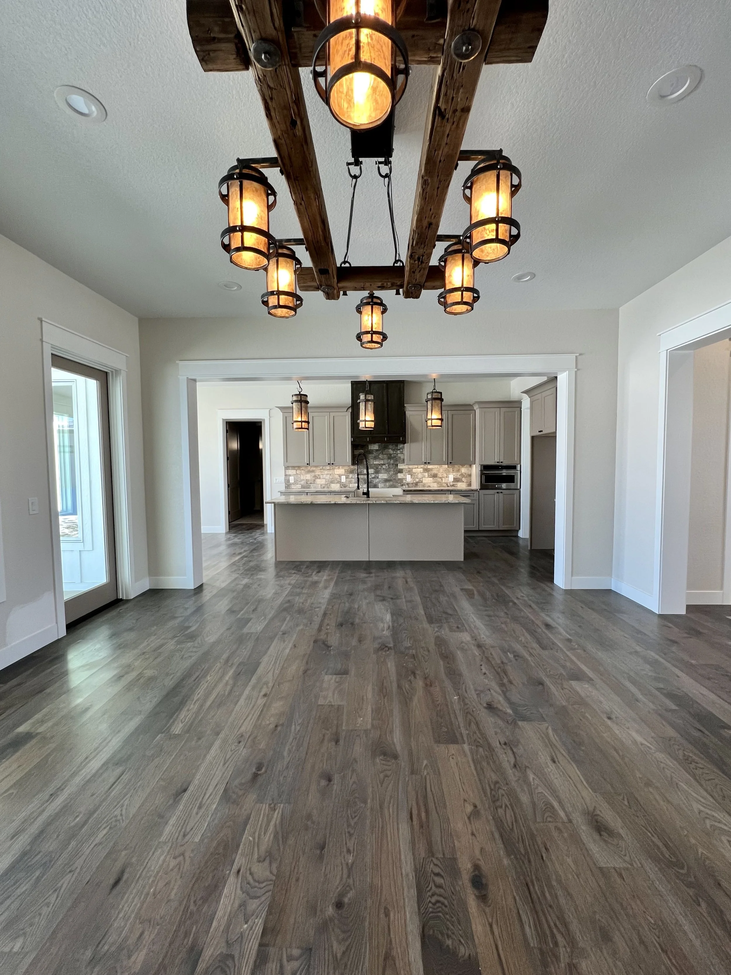 A modern kitchen with an island in the center, pendant lights hanging above, and a stone backsplash. The room has wood flooring and light-colored walls.