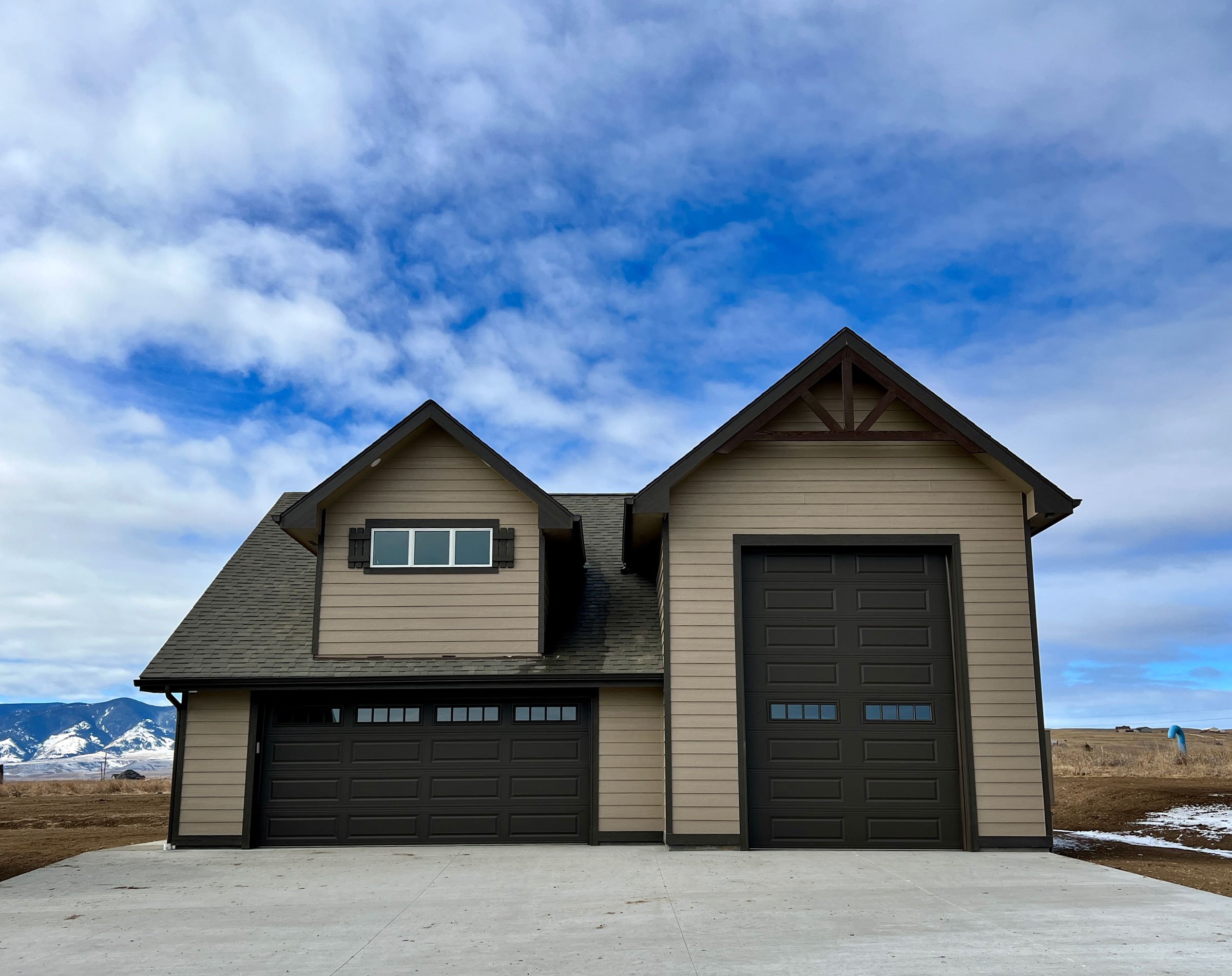 Front view of a new beige house with two black garage doors, two windows, and a peaked roof against a partly cloudy sky with mountains in the background.