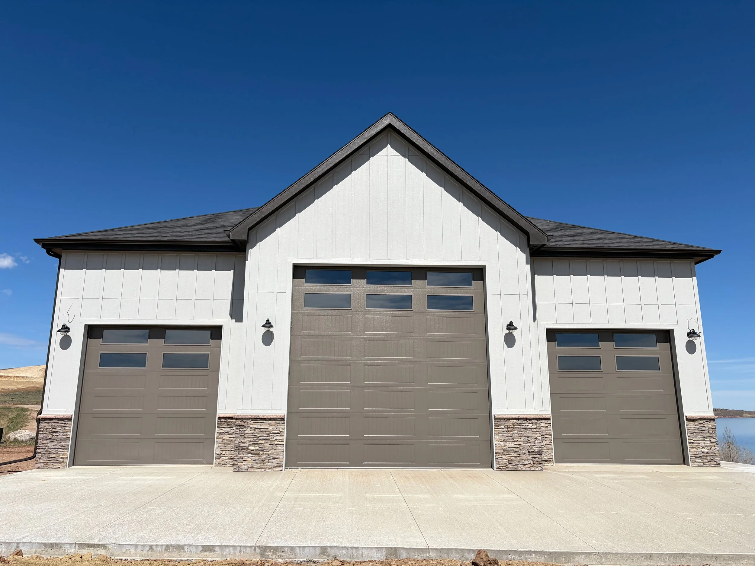 Front view of a modern two-car garage with white vertical siding, stone accent at the bottom, and a dark gray garage door under a gable roof, with black outdoor lights on either side of the garage door. Clear blue sky in the background.