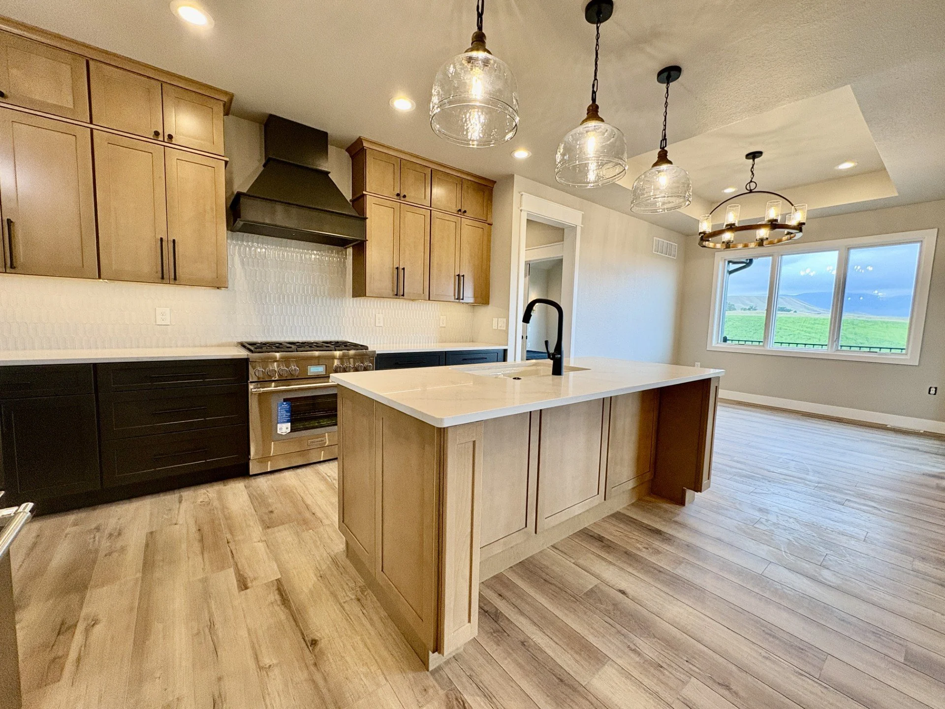 Modern kitchen with a central island, wooden cabinets, stainless steel oven, black and brass pendant lights, large window with a mountain view, wood flooring.