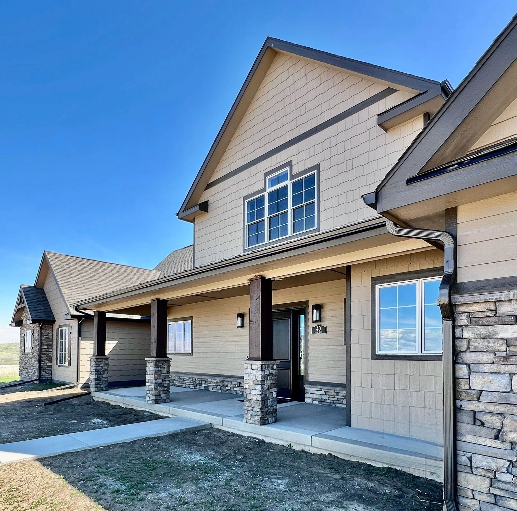 Front view of a new, two-story house with beige siding, stone accents, and large windows under a clear blue sky.