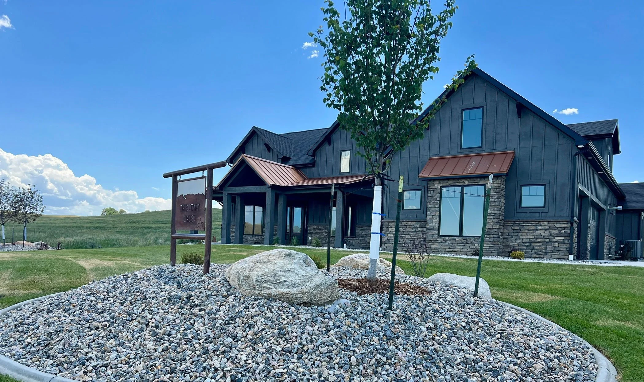 New modern house with black siding and stone accents, several windows, a small tree in the front yard, and a blue sky with scattered clouds.