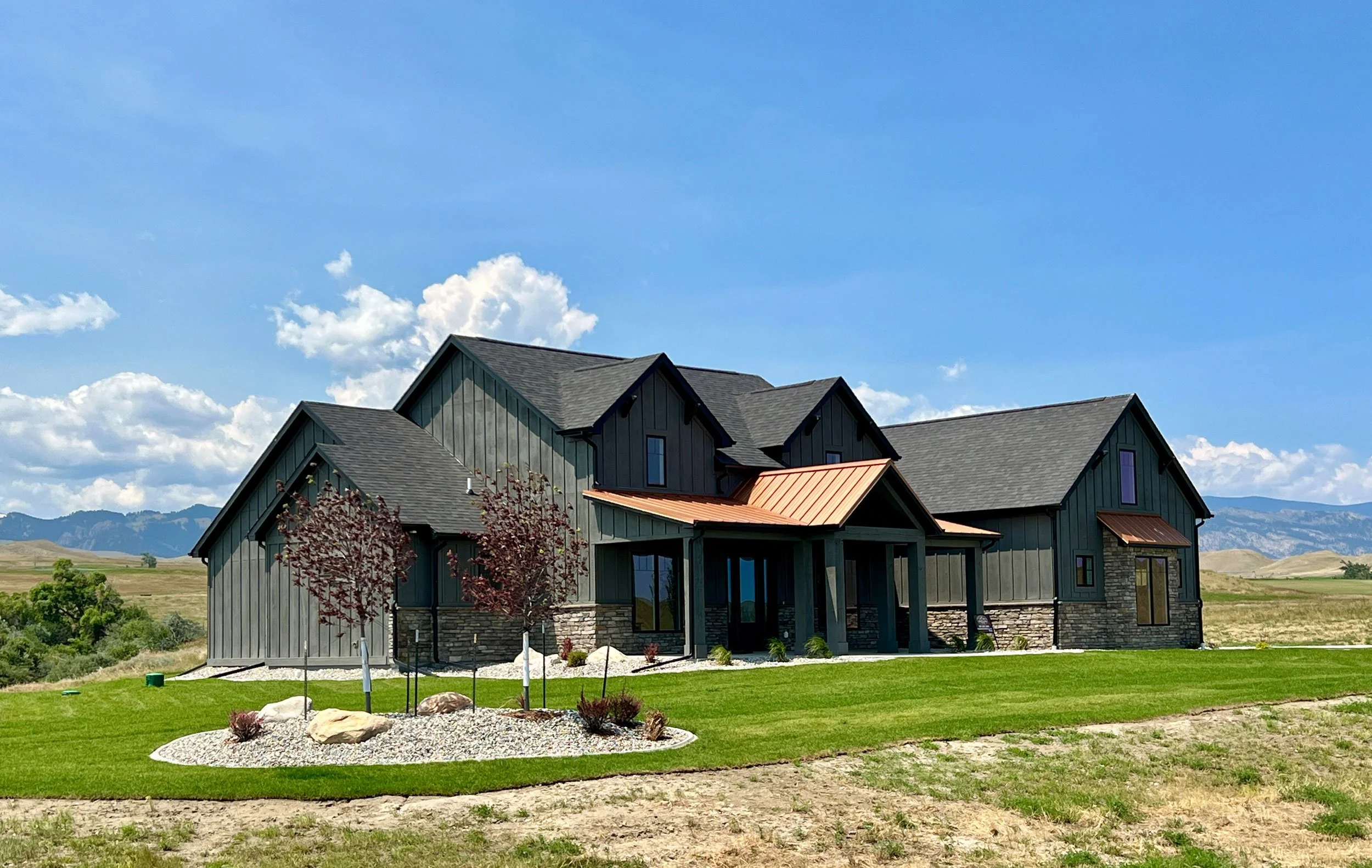 Modern house with dark gray siding, multiple gabled roofs, and a small front porch, set on a lush green lawn with a landscaped garden and trees, against a backdrop of mountains and a partly cloudy sky.