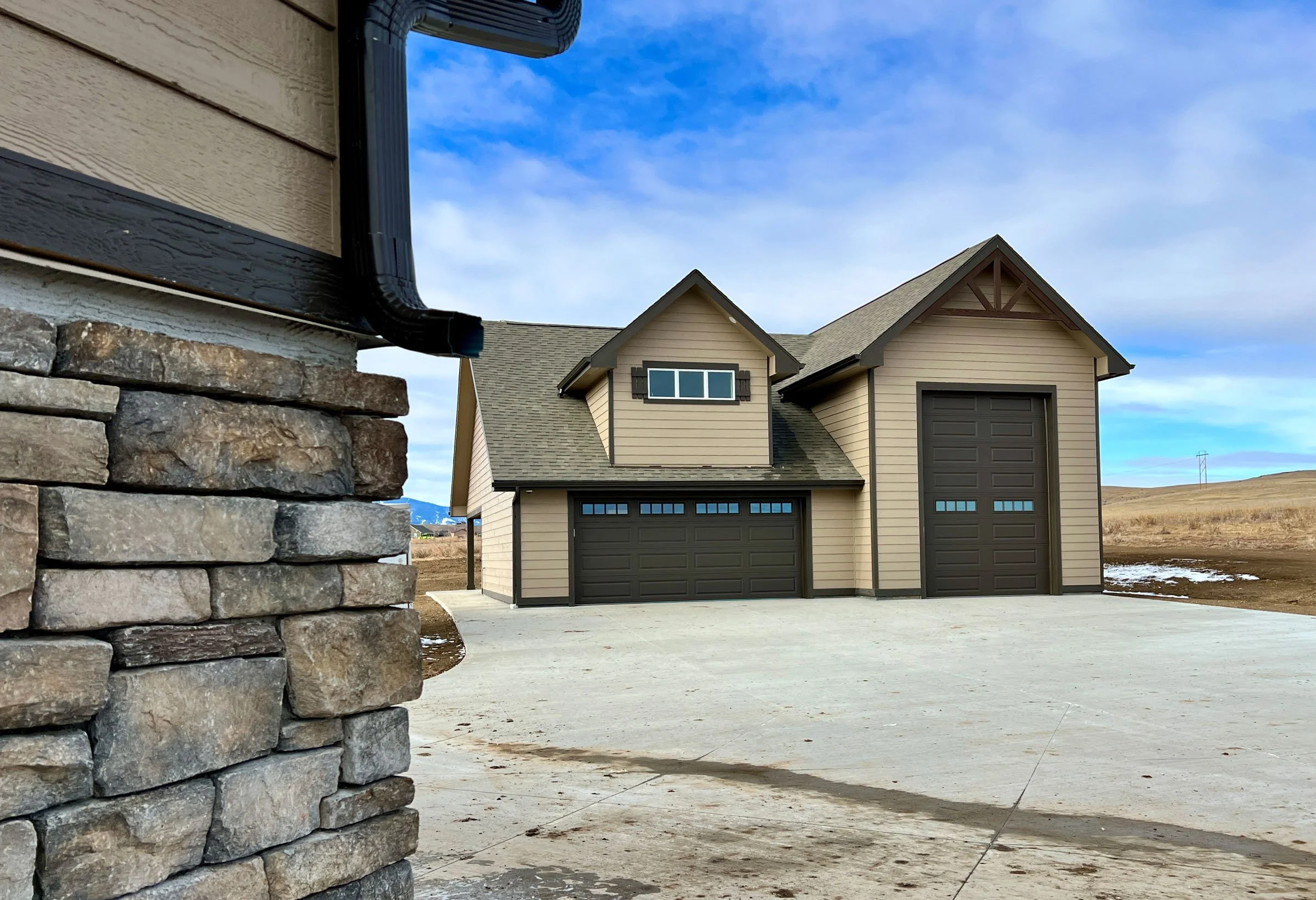 A beige two-story house with a dark gray garage door, black window shutters, and a pitched roof. A stone wall and a black gutter are in the foreground, and a blue sky with some clouds is above.