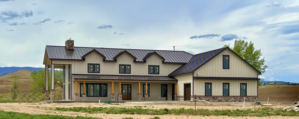 A large two-story house under construction in a rural area with dirt around it, a metal roof, and a mix of siding and stone accents.