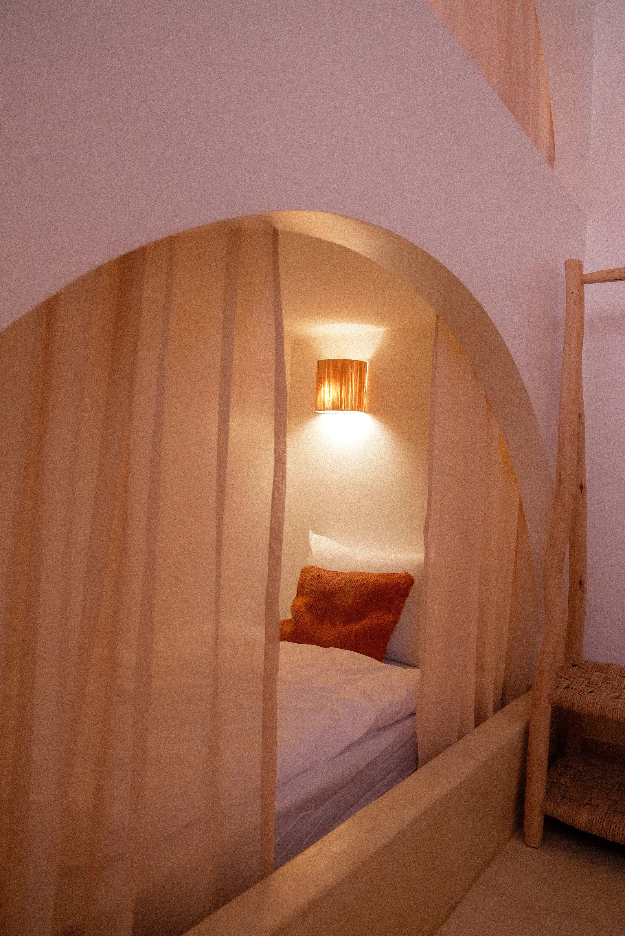 A cozy bedroom with a unique wooden bed frame that has a curved, natural design. The bed is made with white bedding and an orange pillow. There is a warm wall-mounted lamp with a wooden shade illuminating the space, and a woven chair is partly visible on the right side of the image.