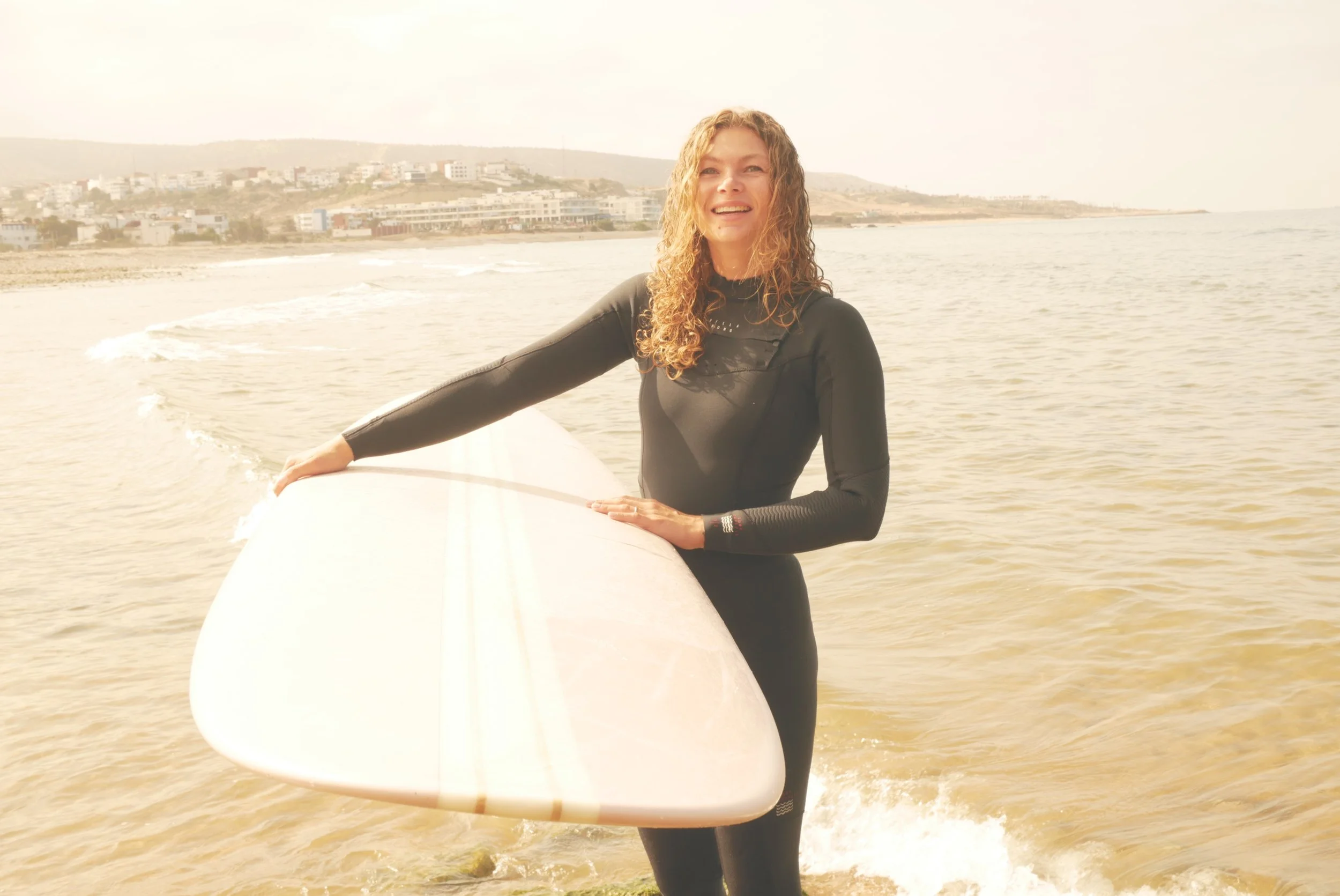 A woman with curly hair in a wetsuit on a surfboard standing in the water at the beach, smiling with buildings on the hillside in the background.