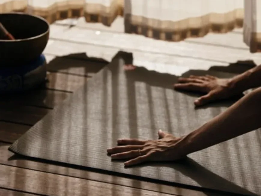 Person practicing yoga on a black mat in an outdoor setting with wooden furniture around.