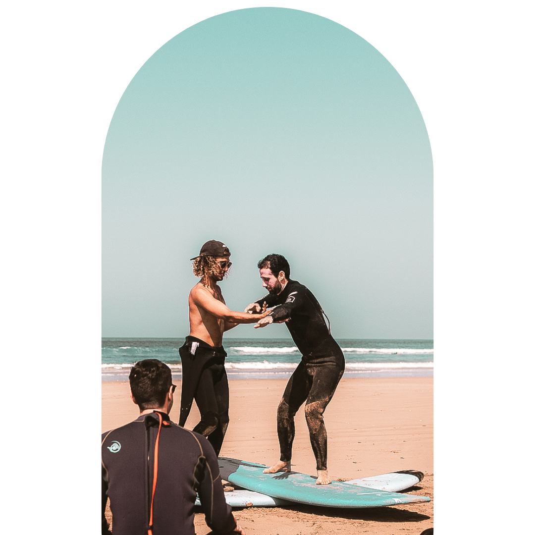 Two surfers, one shirtless with curly hair and the other in a black wetsuit, practicing balance on a surfboard on the beach while an observer watches.