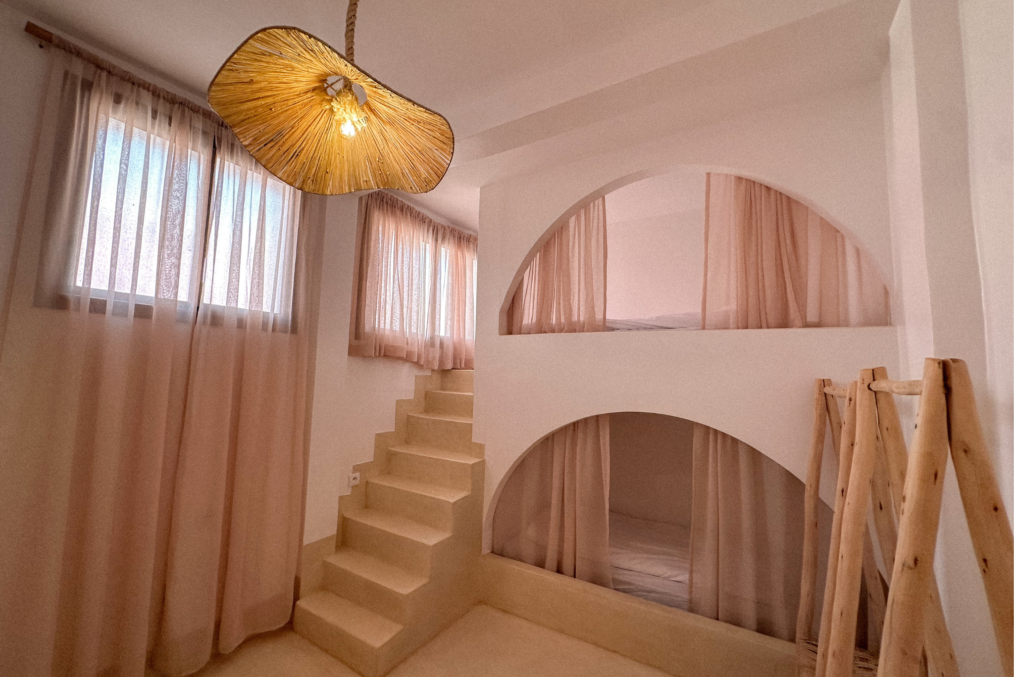 Interior view of a minimalist staircase with wooden steps, curved wooden railing, and sheer pink curtains covering windows, illuminated by a statement light fixture.