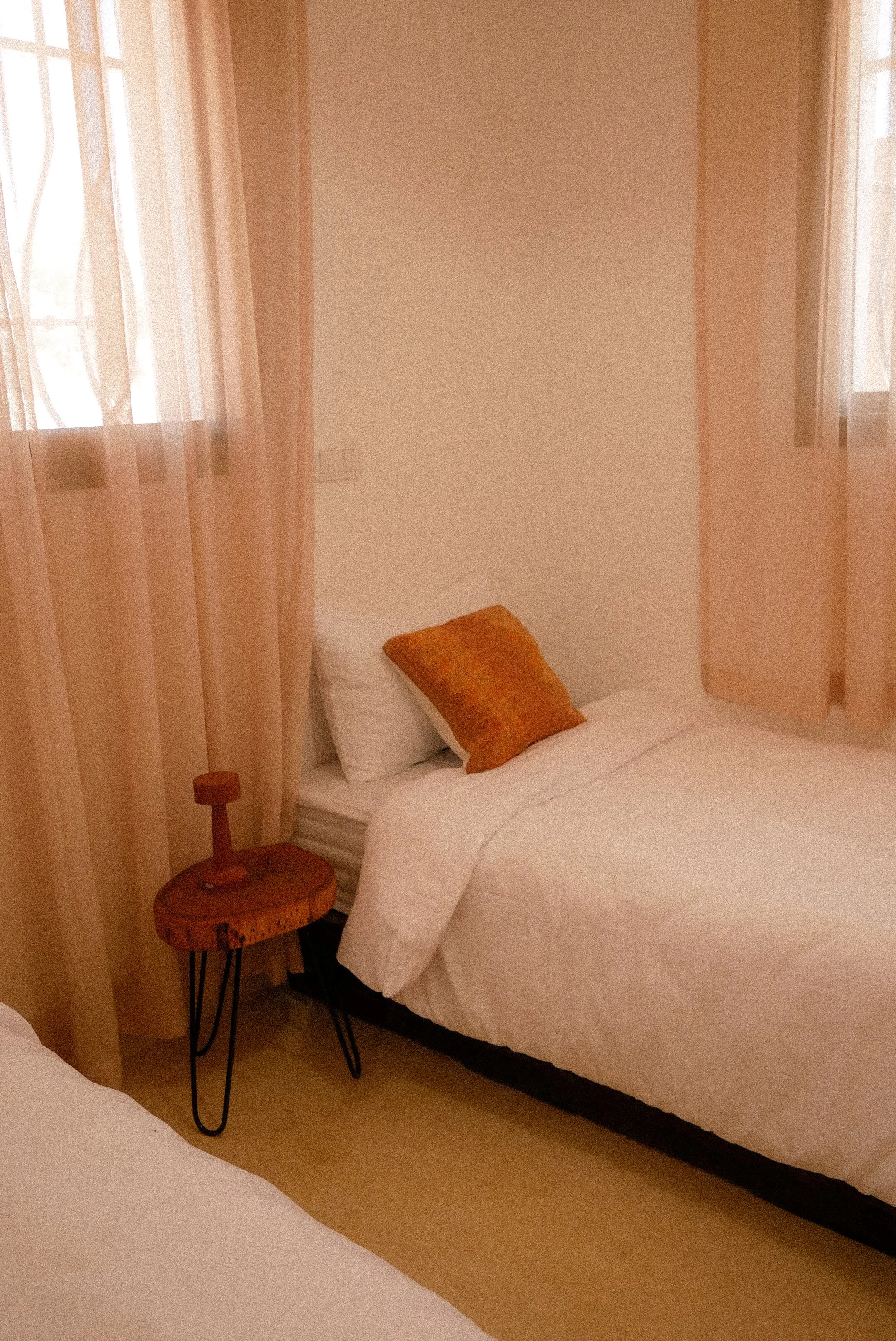 A cozy bedroom with a bed covered in white linens and an orange pillow. Next to the bed is a small wooden side table with black hairpin legs and a modern lamp. The room has sheer peach curtains on the windows, soft natural light entering, and light-colored walls.