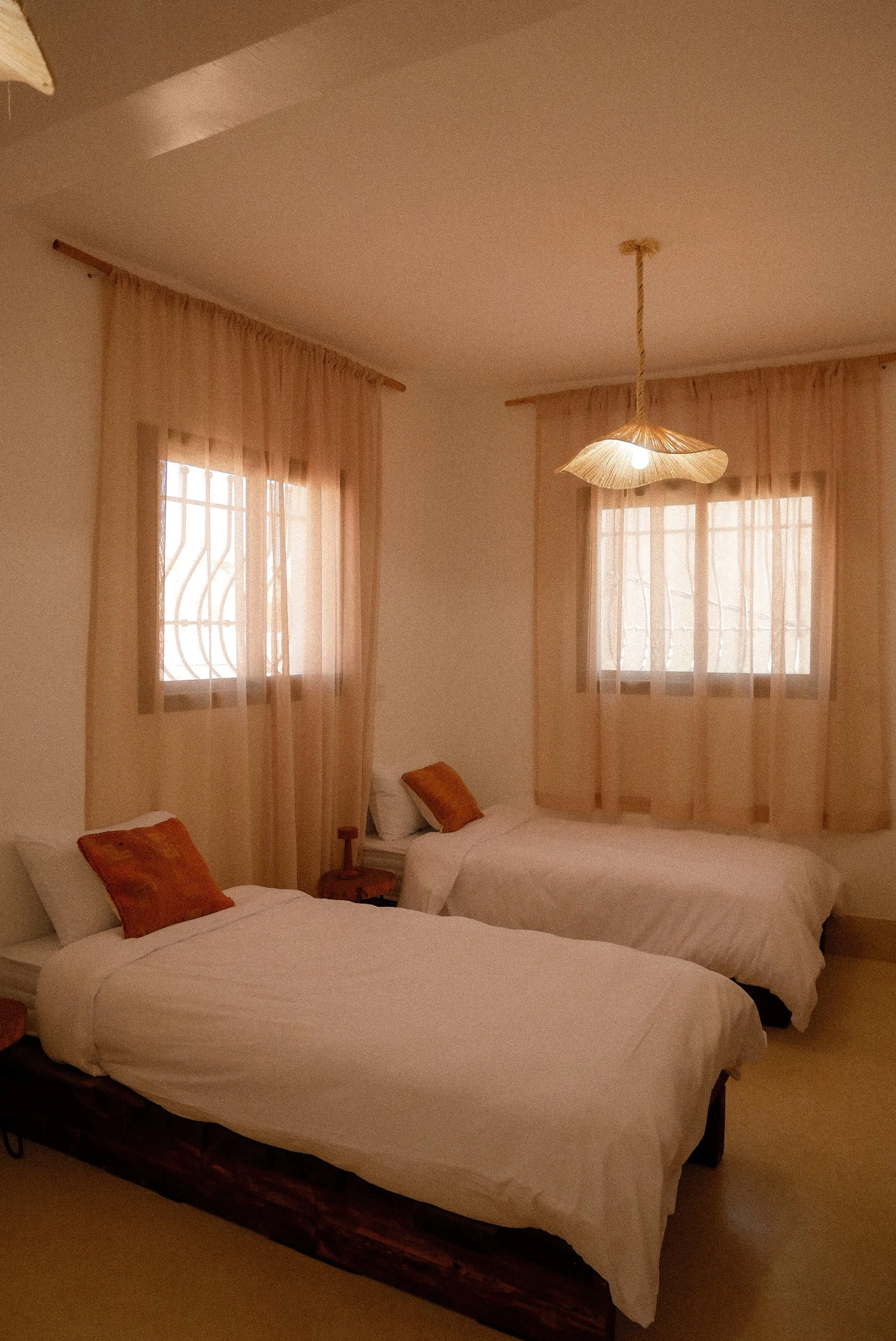 A bedroom with two beds, each with white linens and orange pillows, a bedside table between them, and two windows with sheer beige curtains. A chandelier hangs from the ceiling.