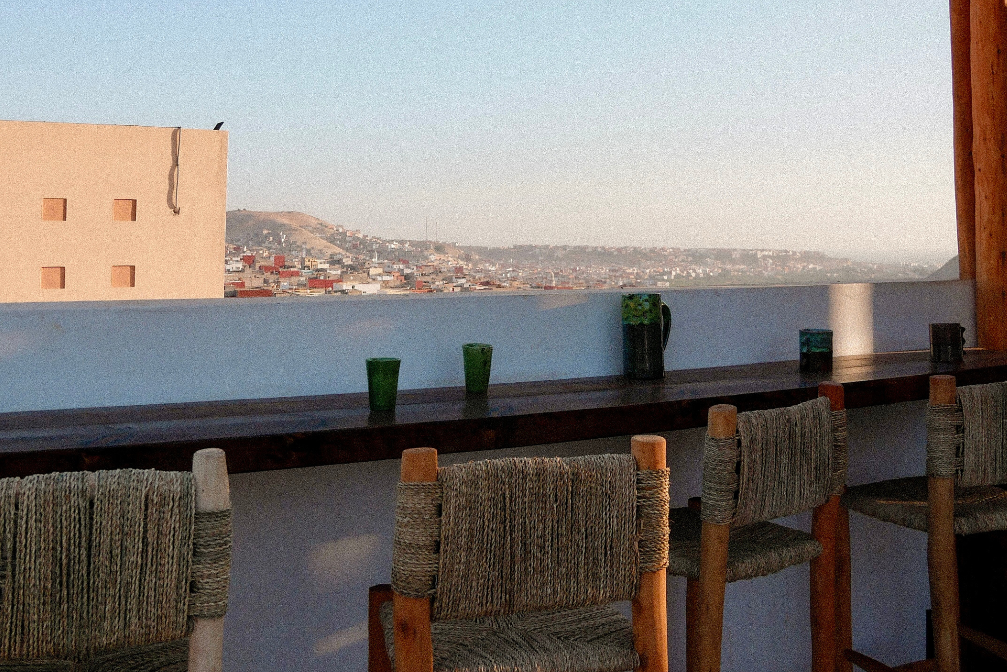 View of a city with many buildings and hills, seen from a balcony with woven chairs and a wooden table with green glasses and pitchers.