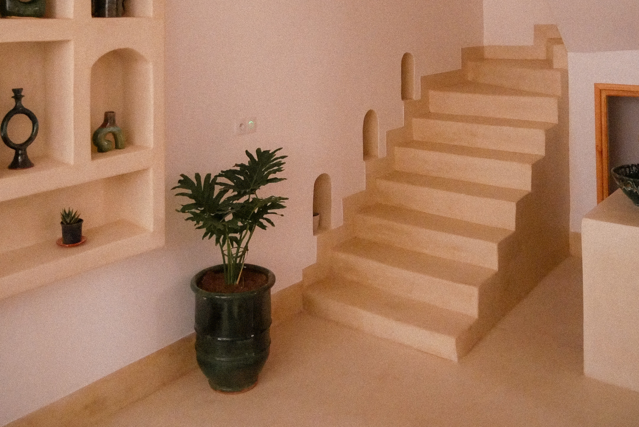 Interior of a house featuring a wooden staircase, built-in wall shelves with decorative vases, and potted plants.