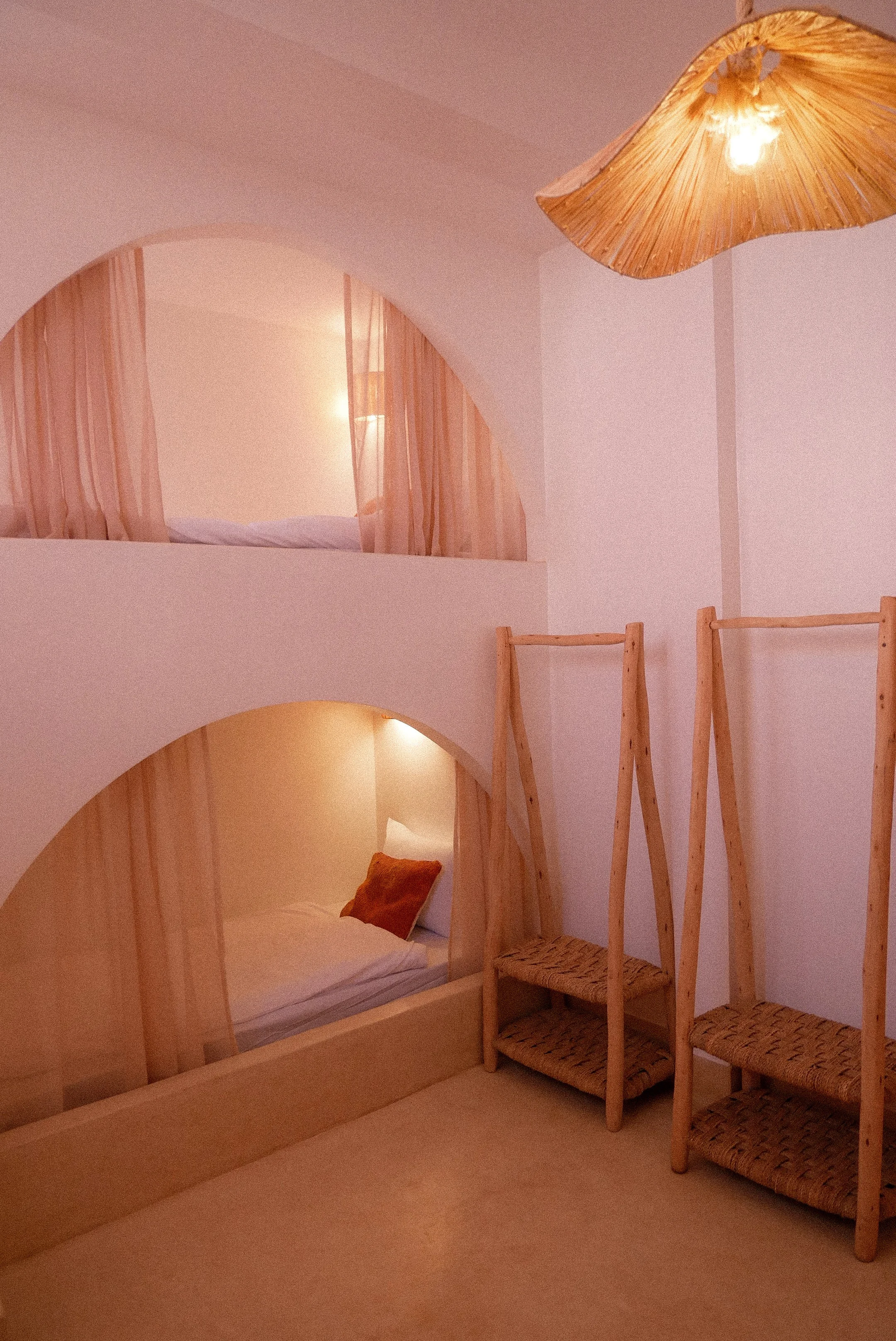 Two minimalist wooden beds with rounded arches, one with beige curtains and the other with a small pillow, located in a room with soft lighting and warm-colored walls, with three woven wooden chairs beside them and a pendant light fixture overhead.