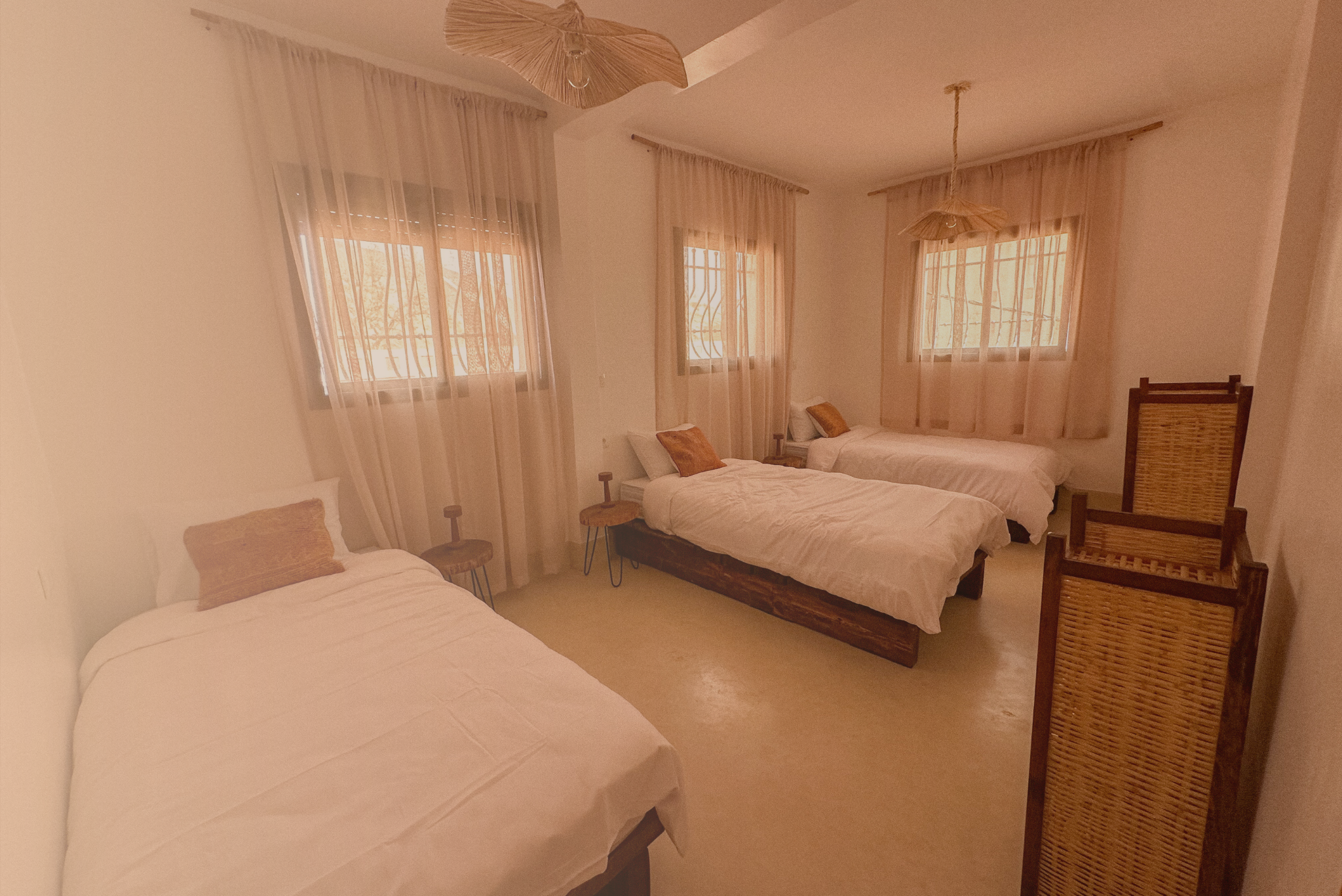 A bedroom with three beds, each with white bedding and brown pillows. The room has three windows with sheer curtains, a rattan ceiling light fixture, a wooden chair, and small bedside tables.