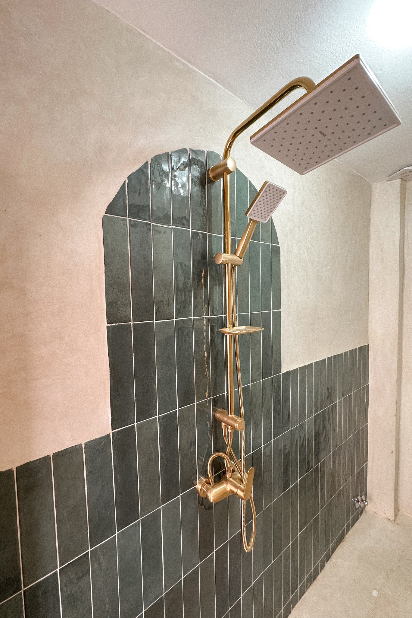 A modern shower with black tiled walls, a gold shower head, and matching fixtures. The shower has a rectangular rain shower head and a handheld shower head, both mounted on the wall. The wall above the tiles is painted in a light beige color.