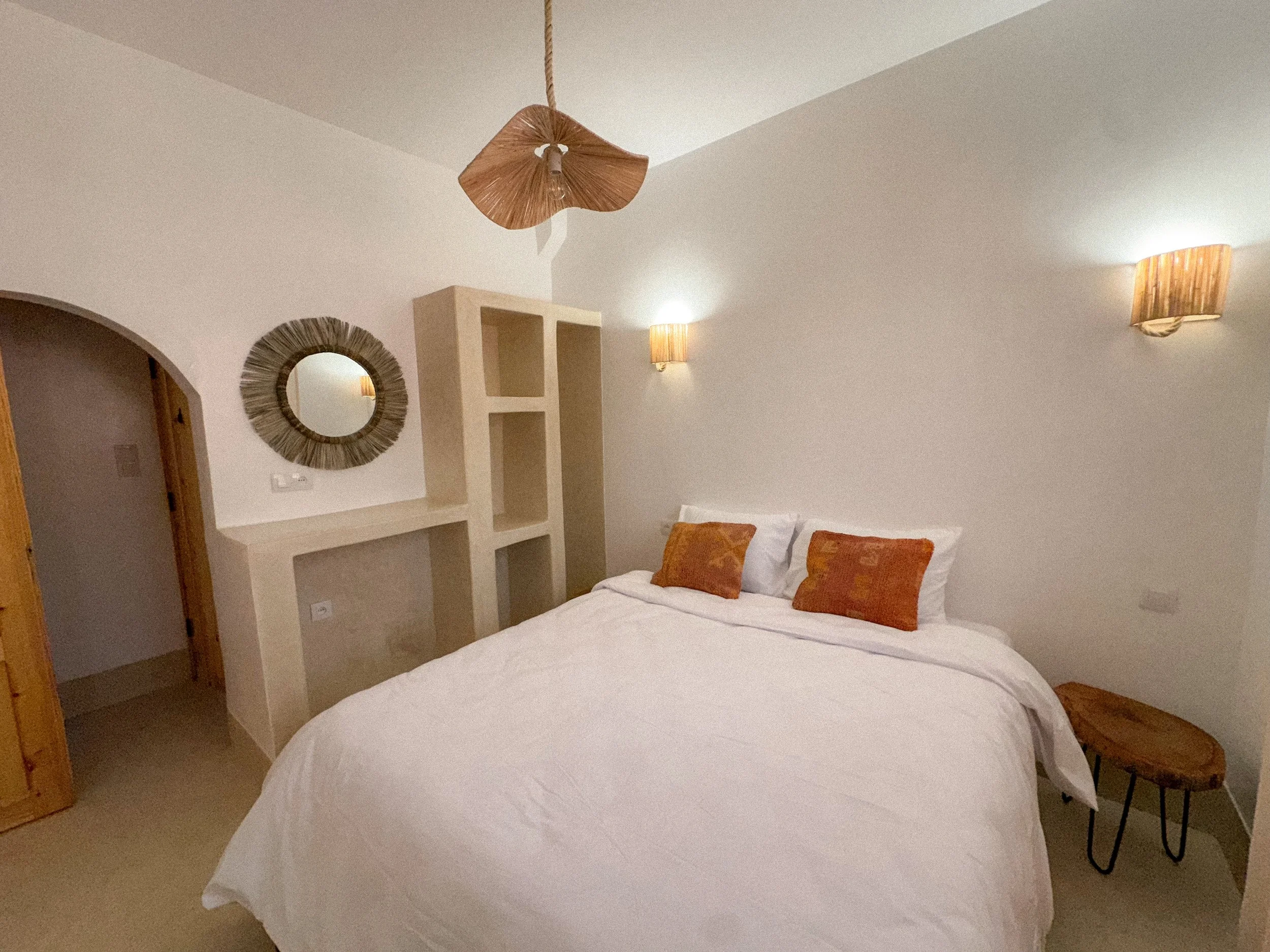 A bedroom with white sheets and pillows on a bed, two orange patterned throw pillows, beige walls with two wall-mounted light fixtures, a round mirror with a textured frame, a built-in shelf, and a ceiling light fixture with a leaf-like design.