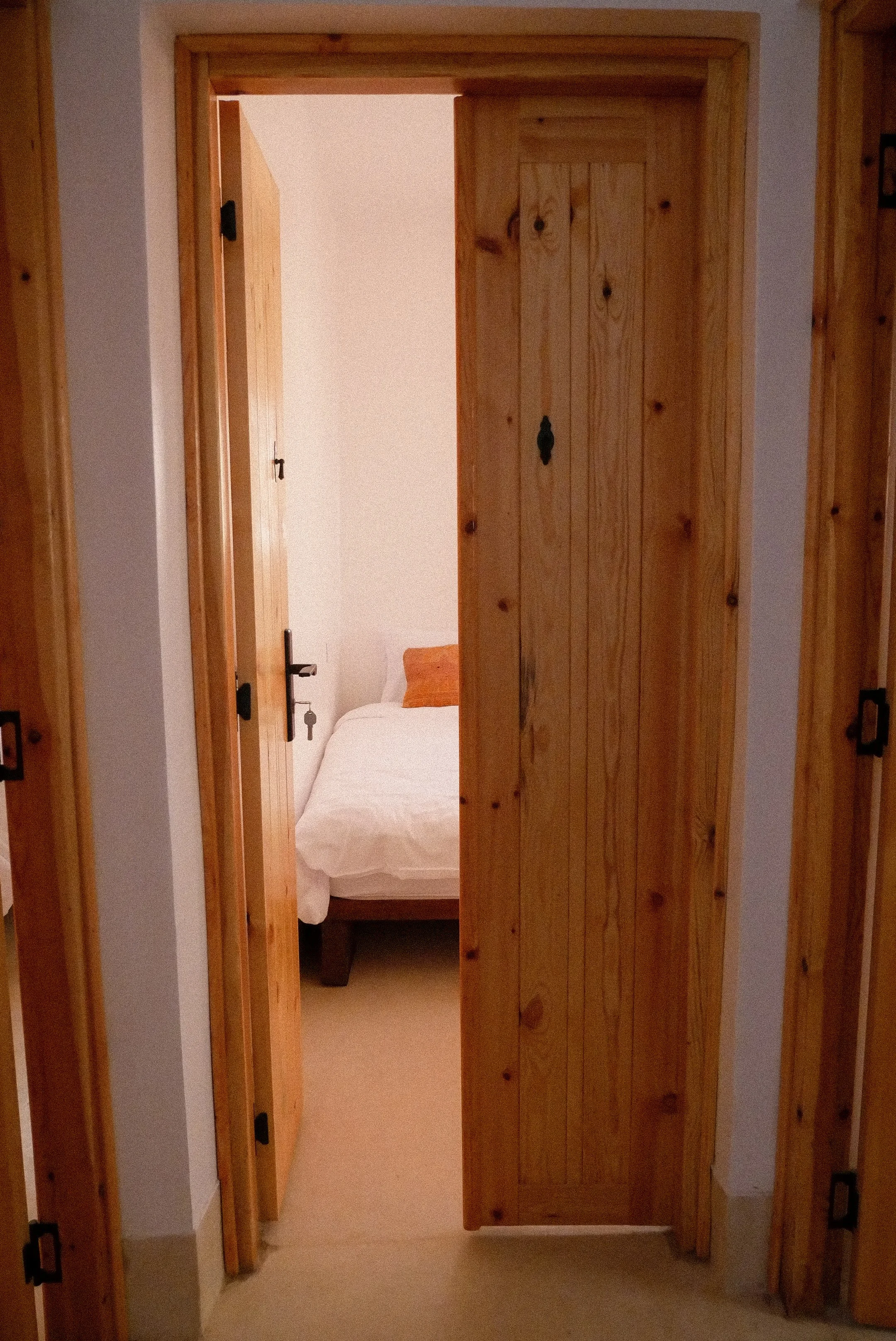 A wooden sliding door partially open, revealing part of a bedroom with white bedding, a wooden bed frame, and an orange pillow.