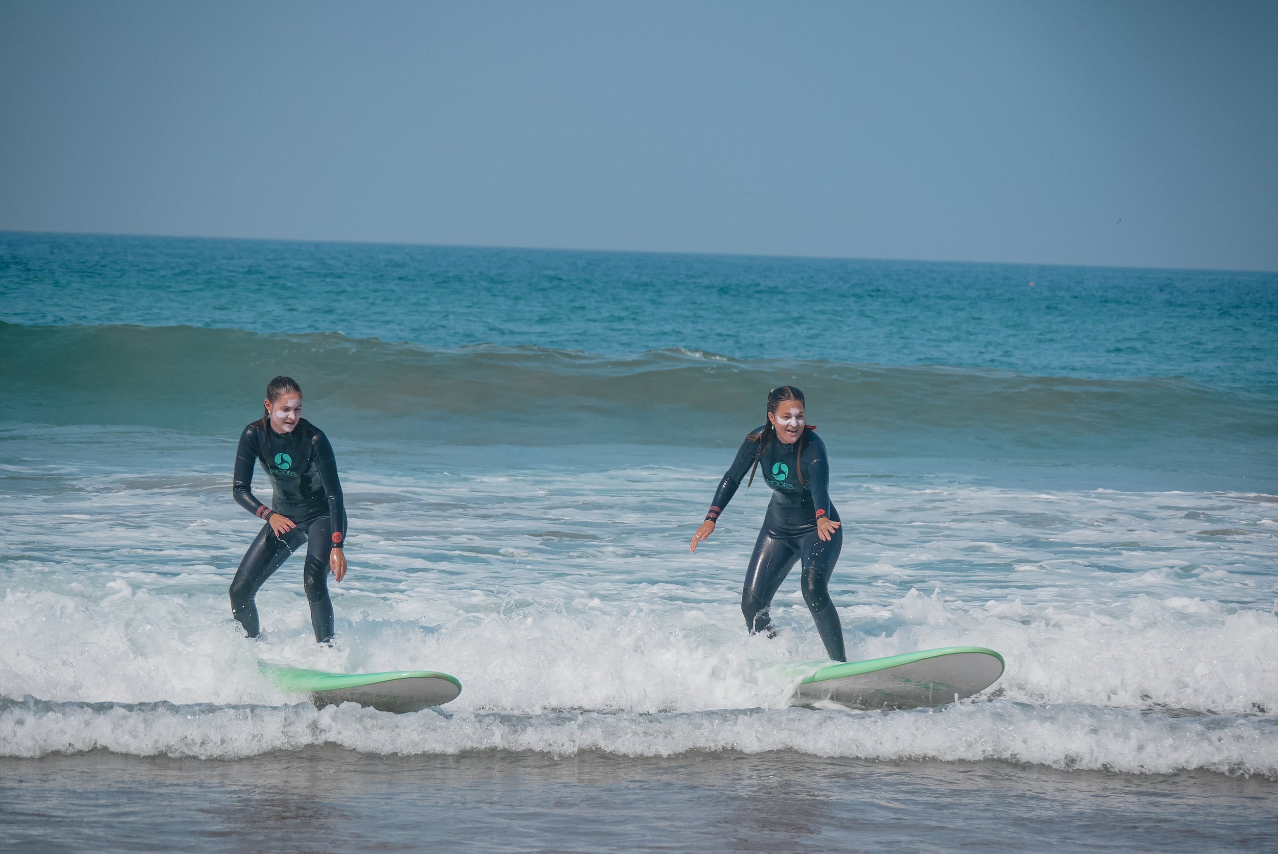 Two young women in wetsuits standing in shallow ocean water, using surfboards on the beach on a sunny day.