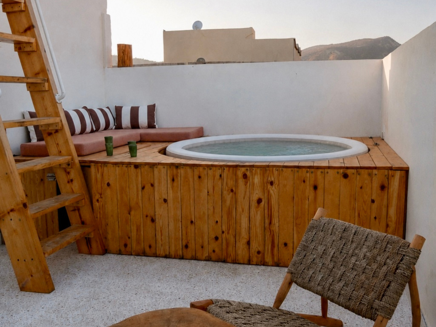 A rooftop patio with a heated jacuzzi in a wooden surround, a cushioned seating area with striped pillows, two green cups, a woven chair, and mountain views in the background.