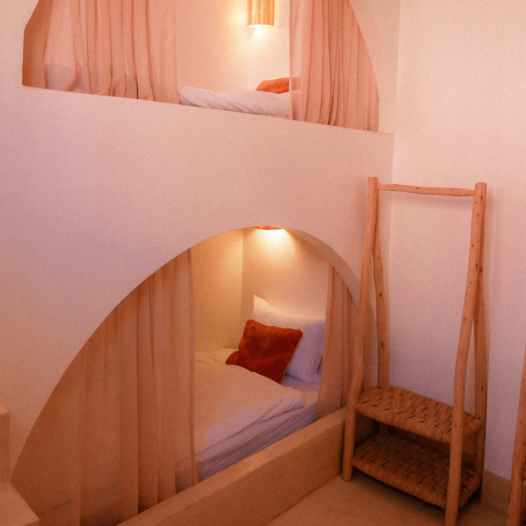 A cozy room with a bunk bed featuring two sleeping areas separated by a circular cutout. The beds have white linens and brown pillows, with warm lighting creating a inviting atmosphere. There is a wooden ladder leaning against the wall for access to the top bed.