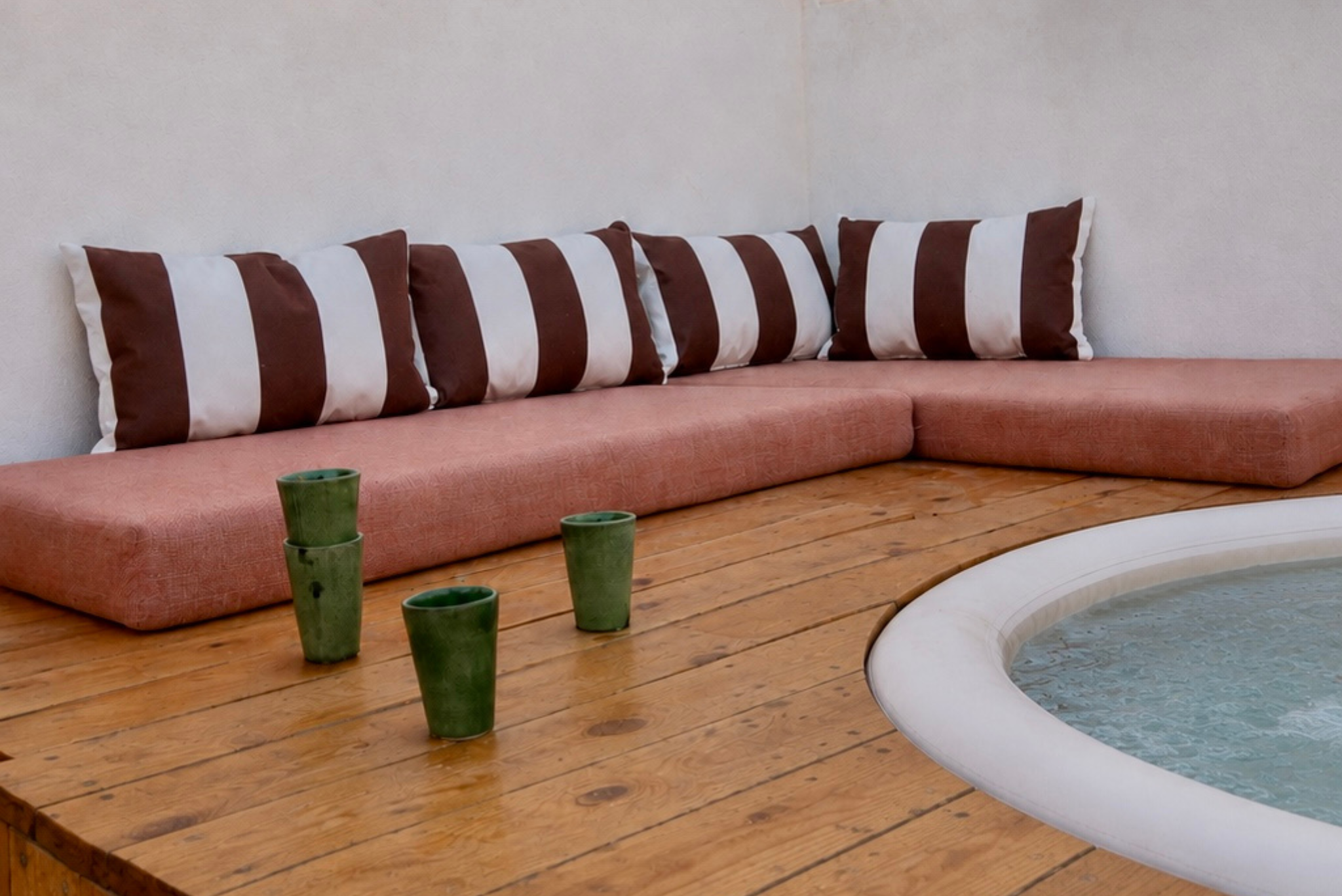 A corner of a room with a wooden deck floor, a built-in peach-colored couch with striped pillows, and a small round hot tub with water, surrounded by four green cups.