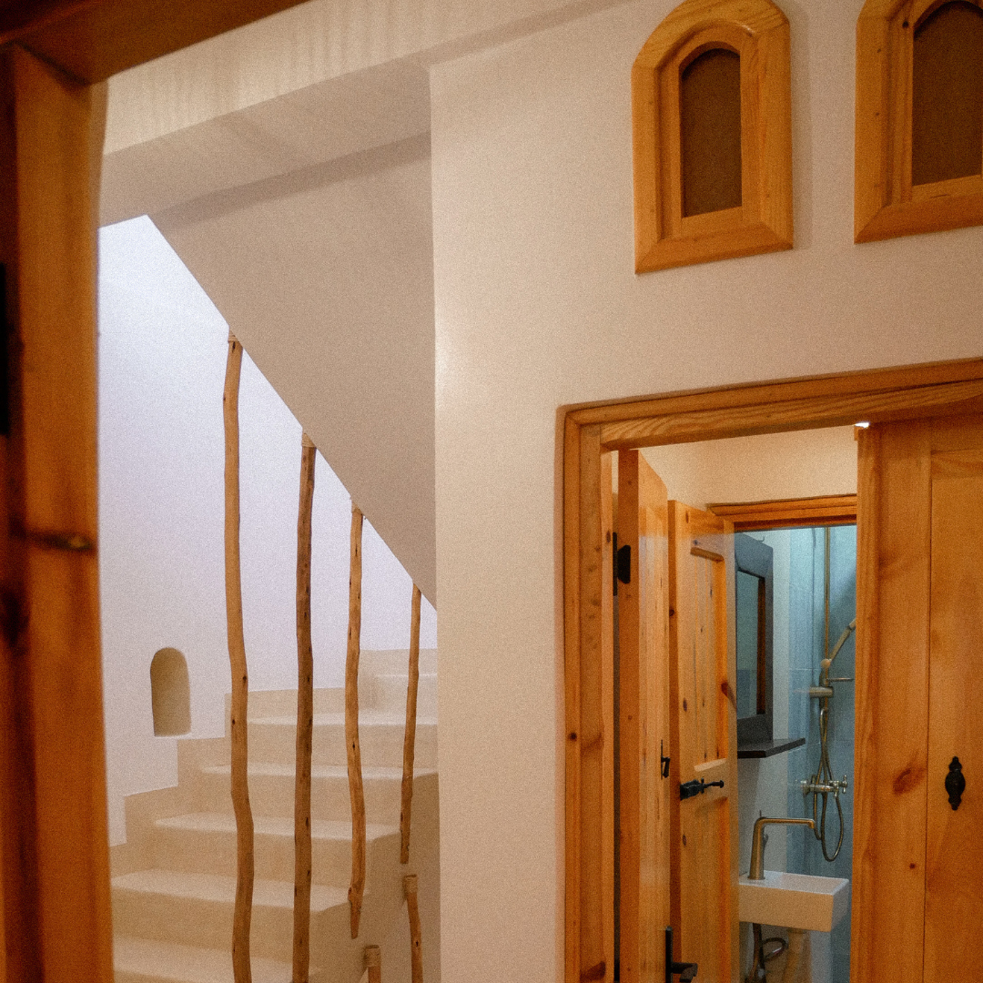 Interior of a rustic home with wooden accents. There are stairs with twig-like wooden balusters, small arched windows with wooden frames, and a doorway leading to a bathroom with a blue wall, a white sink, and a shower.