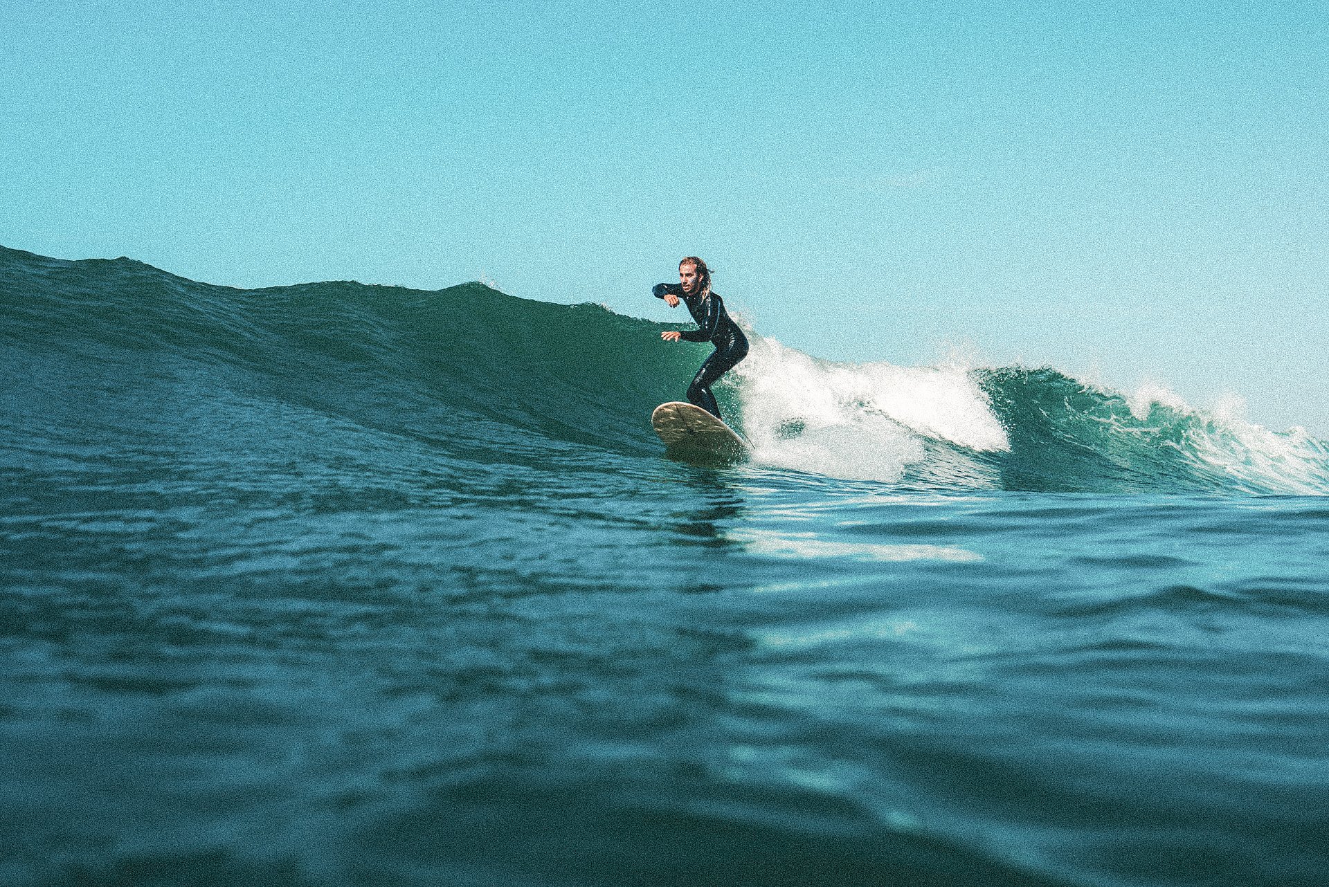 Person surfing on a wave in the ocean, wearing a wetsuit and riding a surfboard under a clear sky.