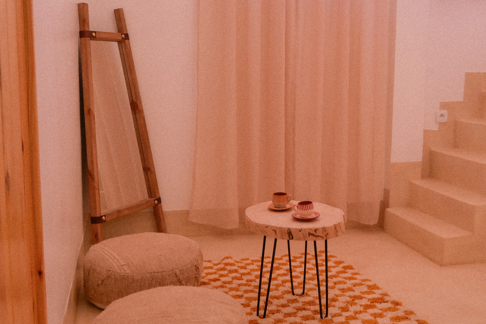 A cozy room with a pink curtain, a beige upholstered bed, a small round wooden table with three ceramic bowls, a tall wooden mirror, and a staircase with a white wall and orange and white geometric patterned rug.