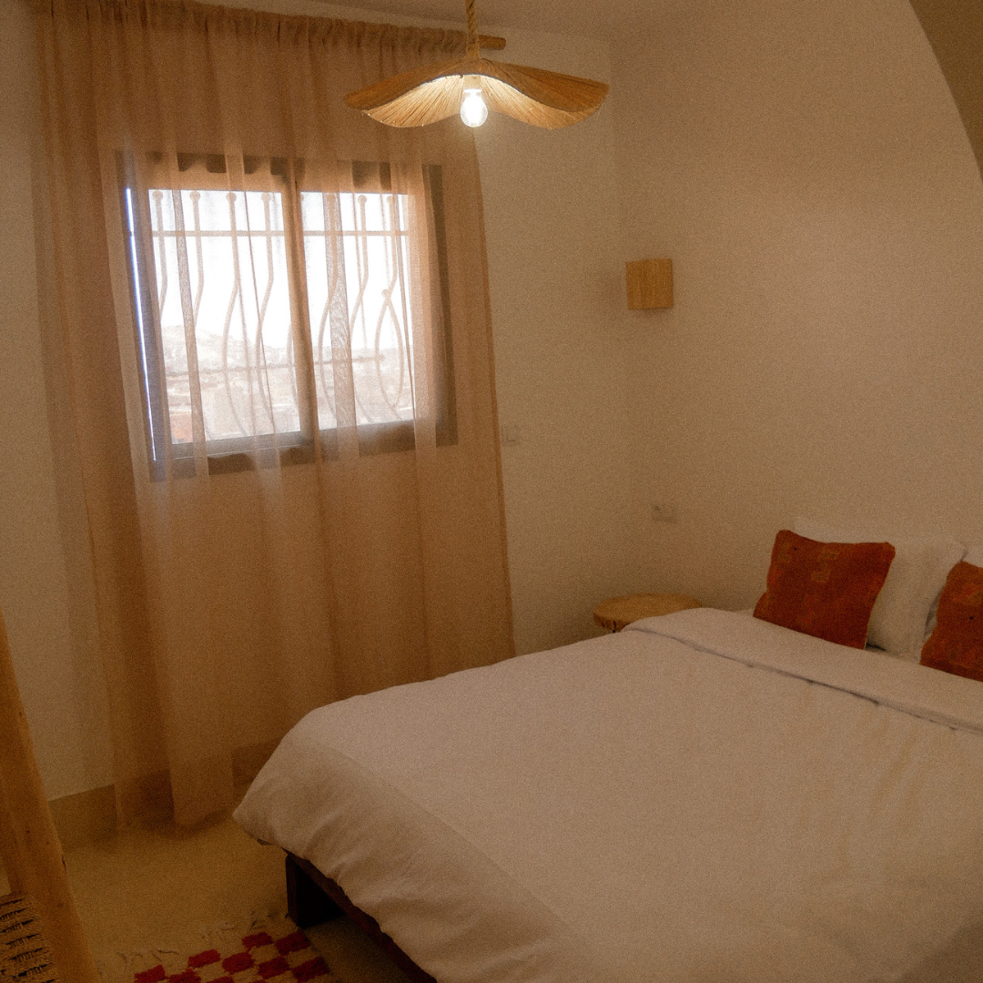 A cozy bedroom with a white bed, orange pillows, a small wooden nightstand, sheer beige curtains, a window with decorative metal bars, and a ceiling fan with leaf-shaped blades.