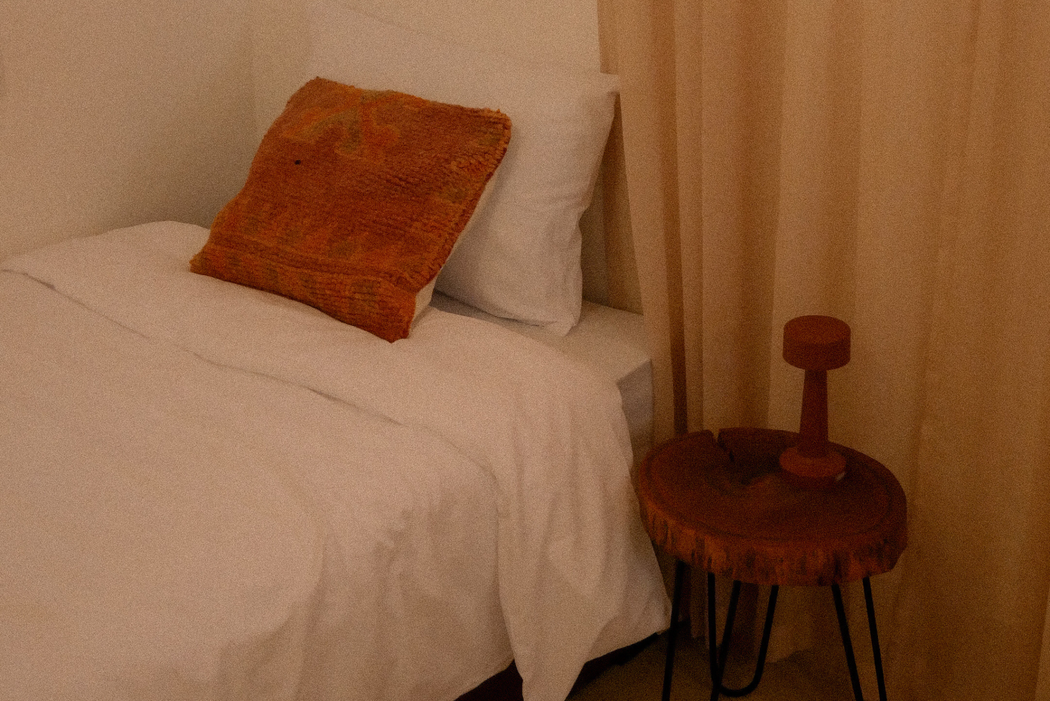 A close-up view of a bed with white bedding and a brown textured pillow. Next to the bed is a small round wooden side table with a short, brown lampshade. The wall behind is plain and light-colored.