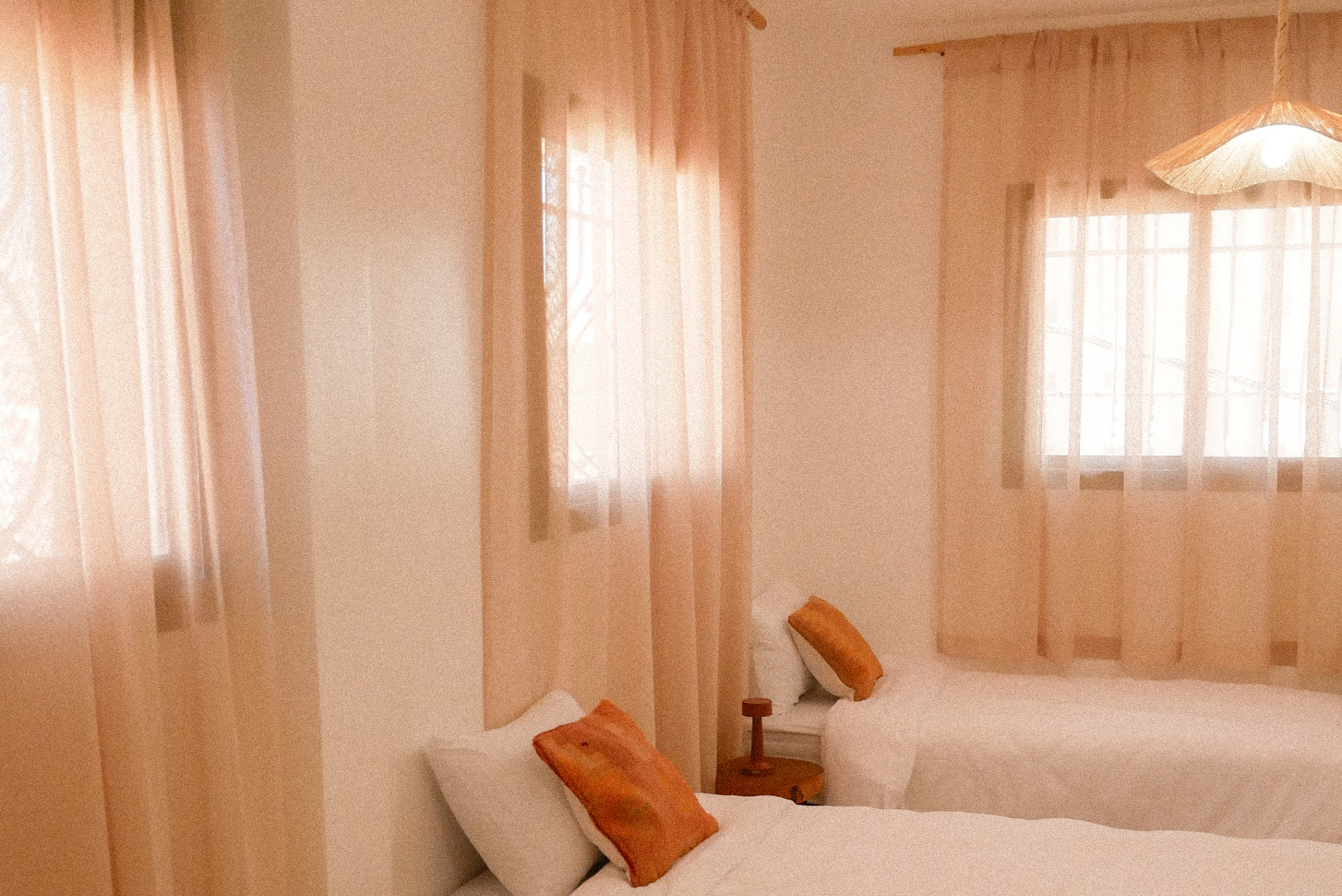 Bedroom with beige walls, wooden pillows on bed, window with sheer curtains, hanging light fixture.