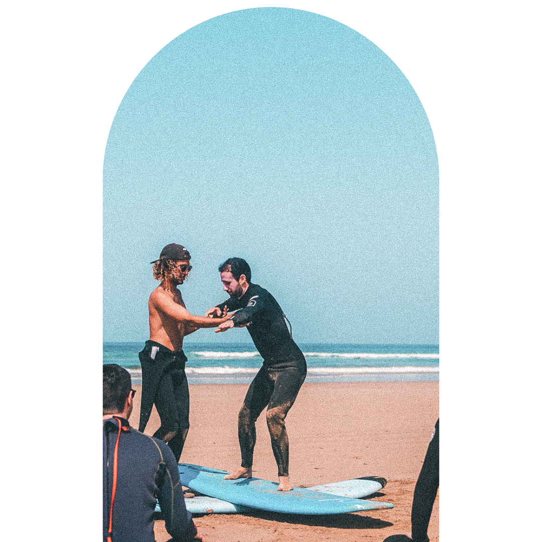 Two men on a beach, one in a wetsuit, teaching the other how to surf, with friends watching nearby.