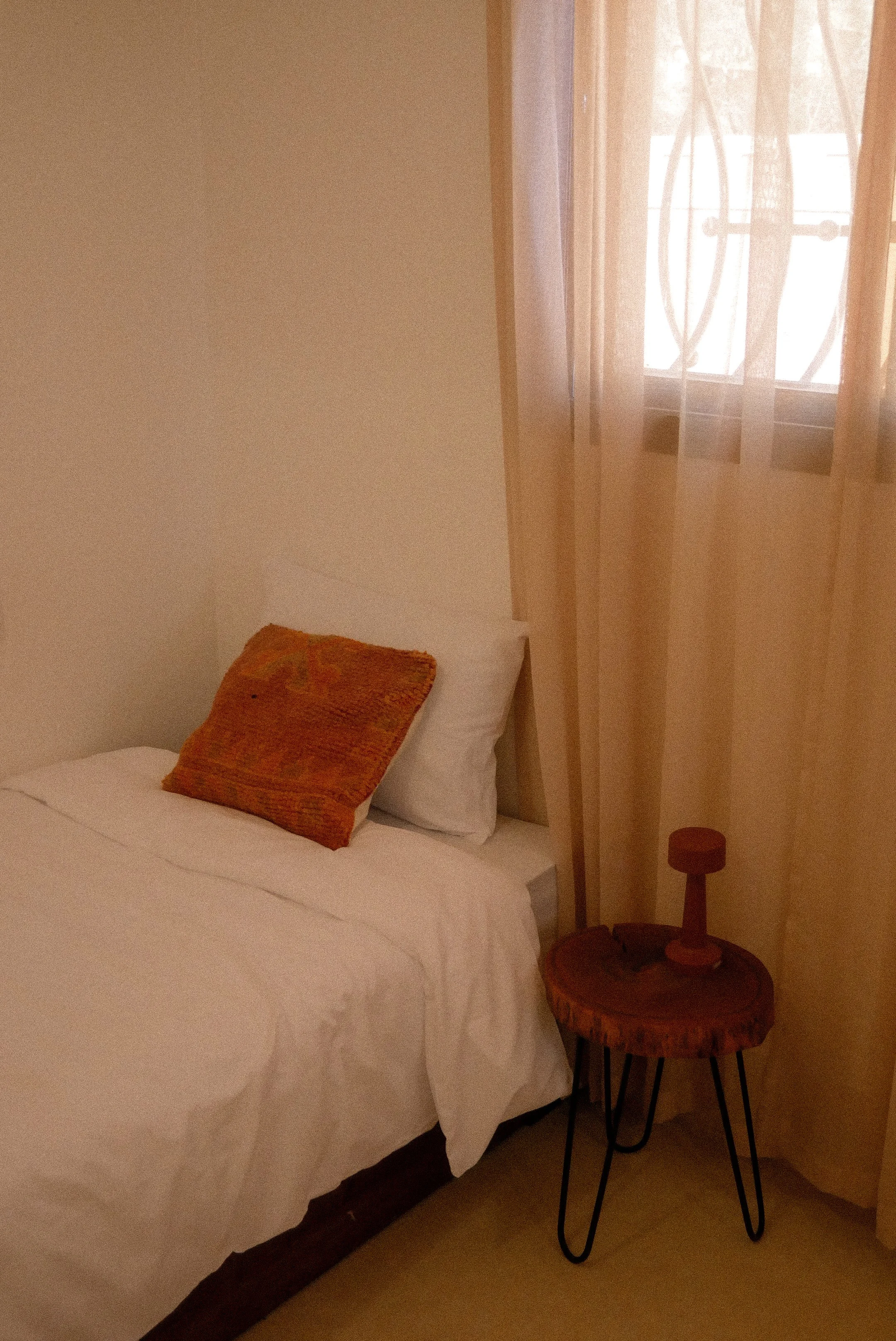 A bedroom with a bed covered in white bedding, a small wooden nightstand, a window with sheer beige curtains, a pillow on the bed, and a small round side table with a wooden structure and black metal legs.