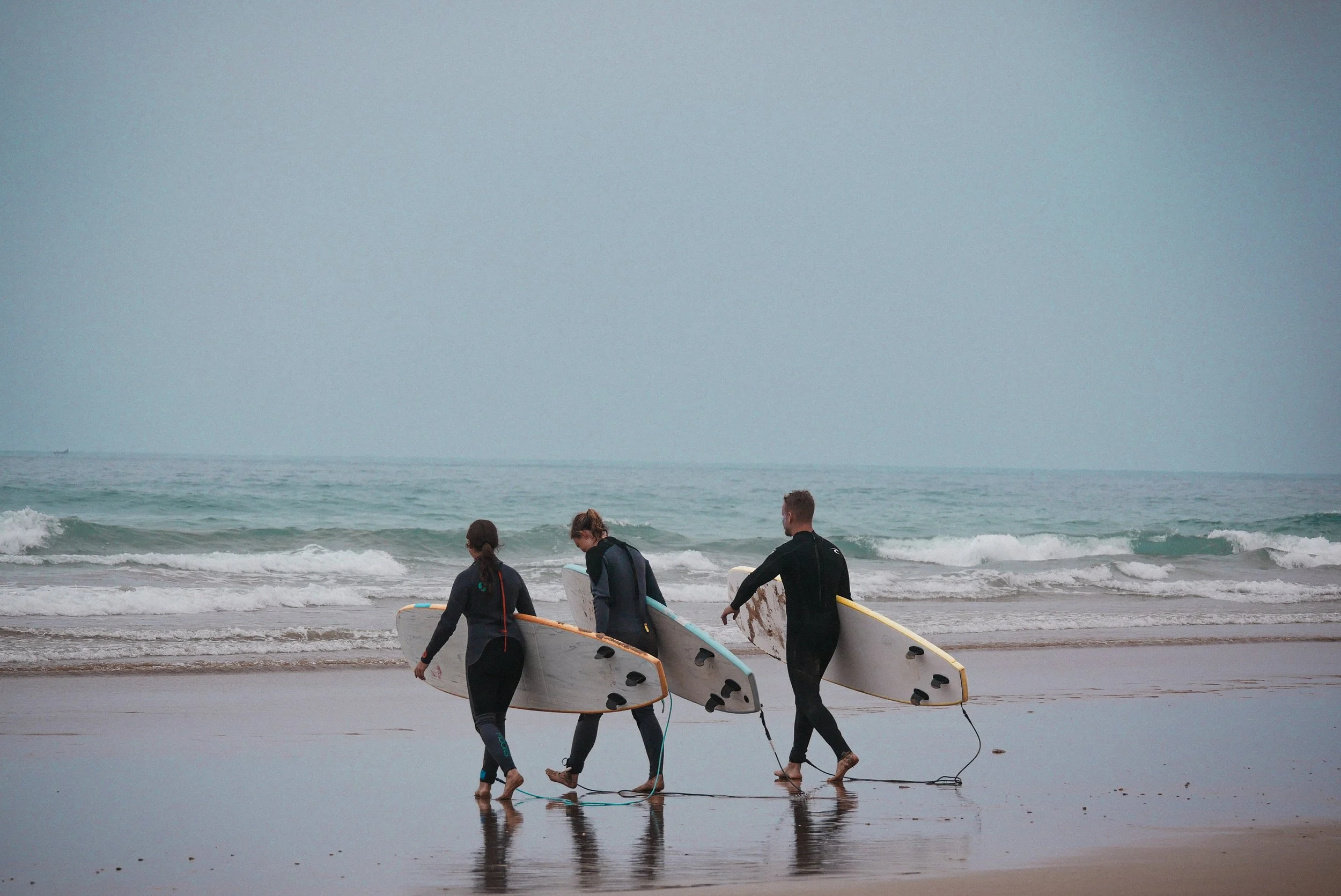Three surfers carrying surfboards along the shoreline, with the ocean and a cloudy sky in the background.