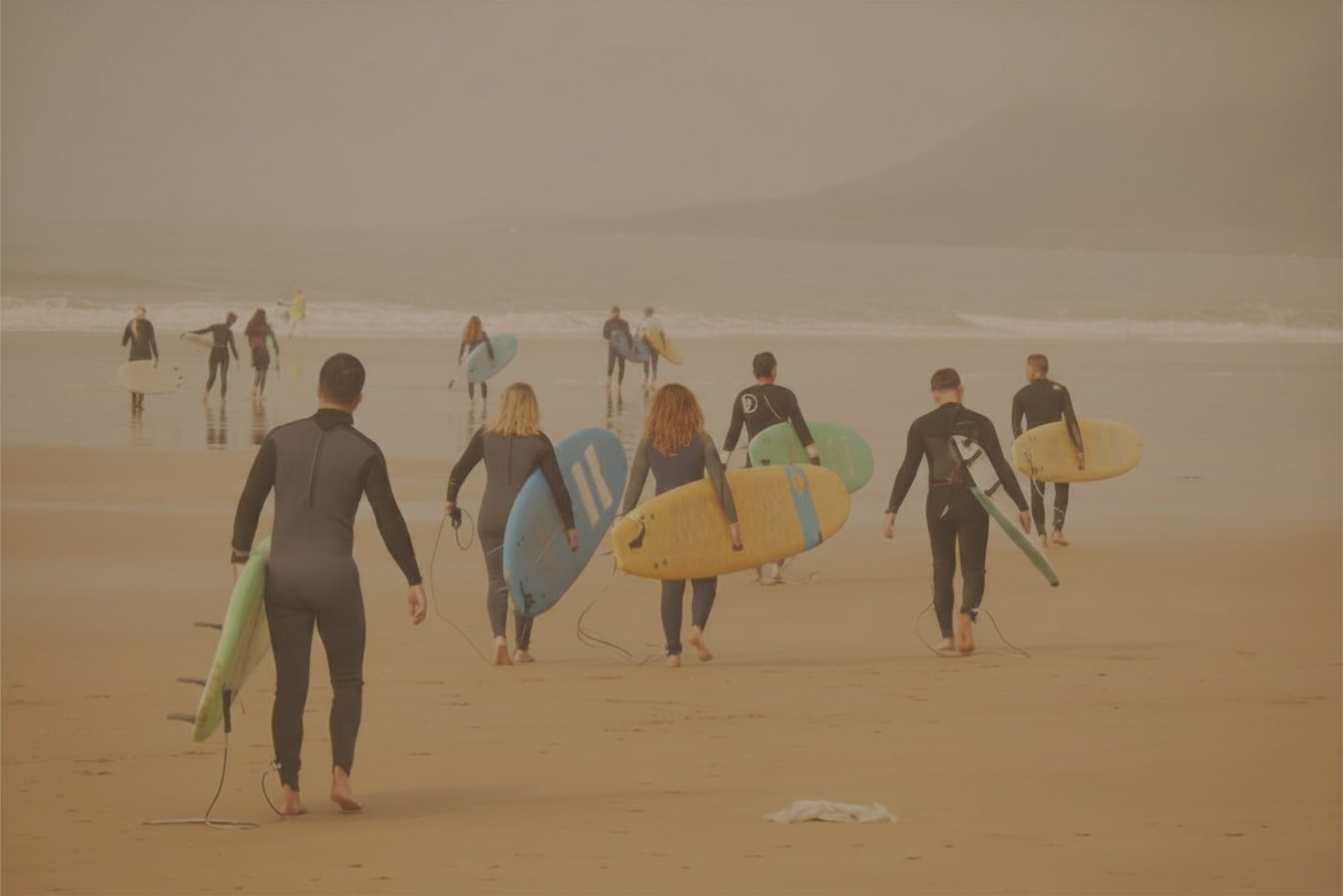 People wearing wetsuits and carrying surfboards walking toward the ocean on a sandy beach.