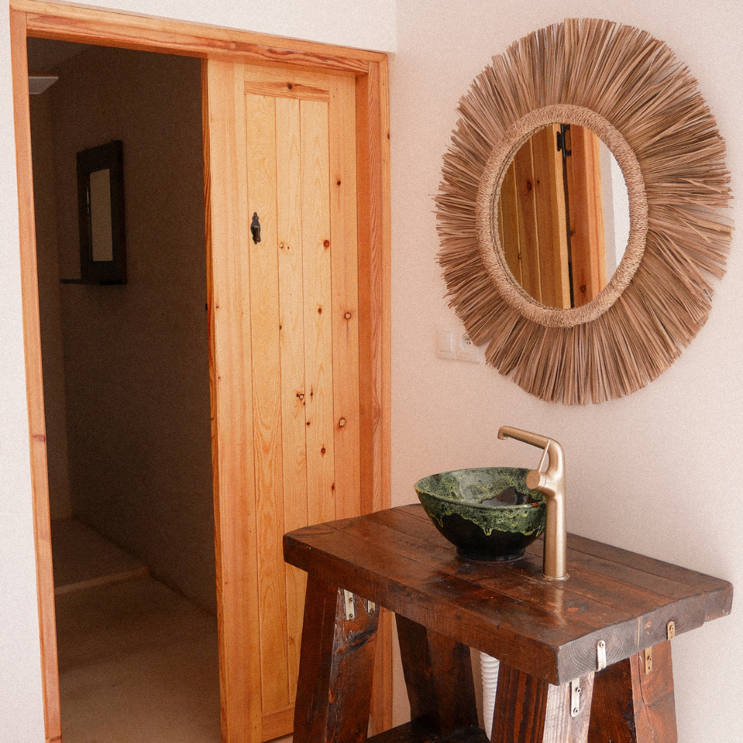 A rustic bathroom with a wooden vanity, a green stone sink, and a gold faucet. A large round decorative mirror with a sunburst frame hangs on the wall.