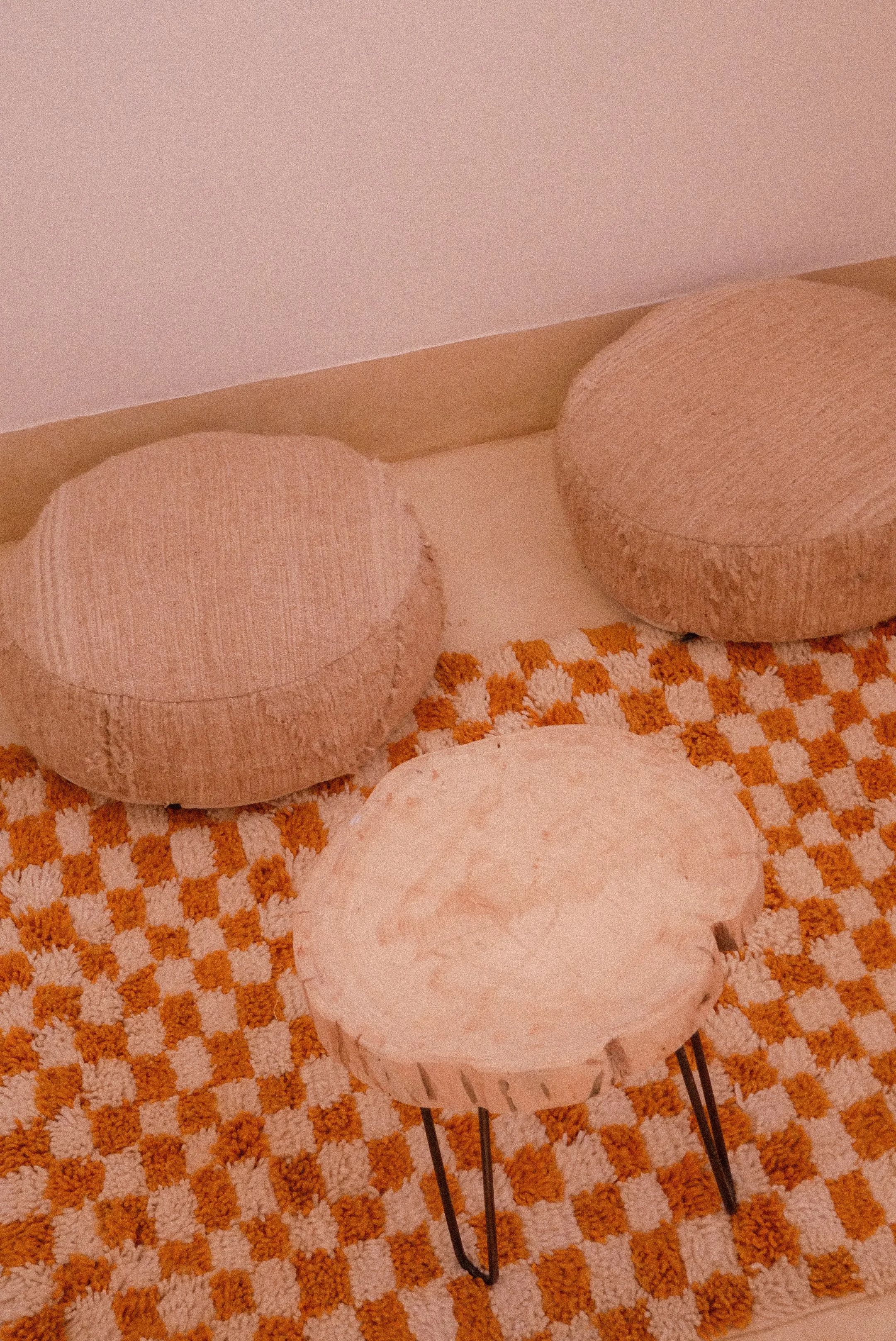 A small round white marble table with black legs, placed on an orange and white checkered rug, with beige upholstered ottomans behind it and a plain wall in the background.