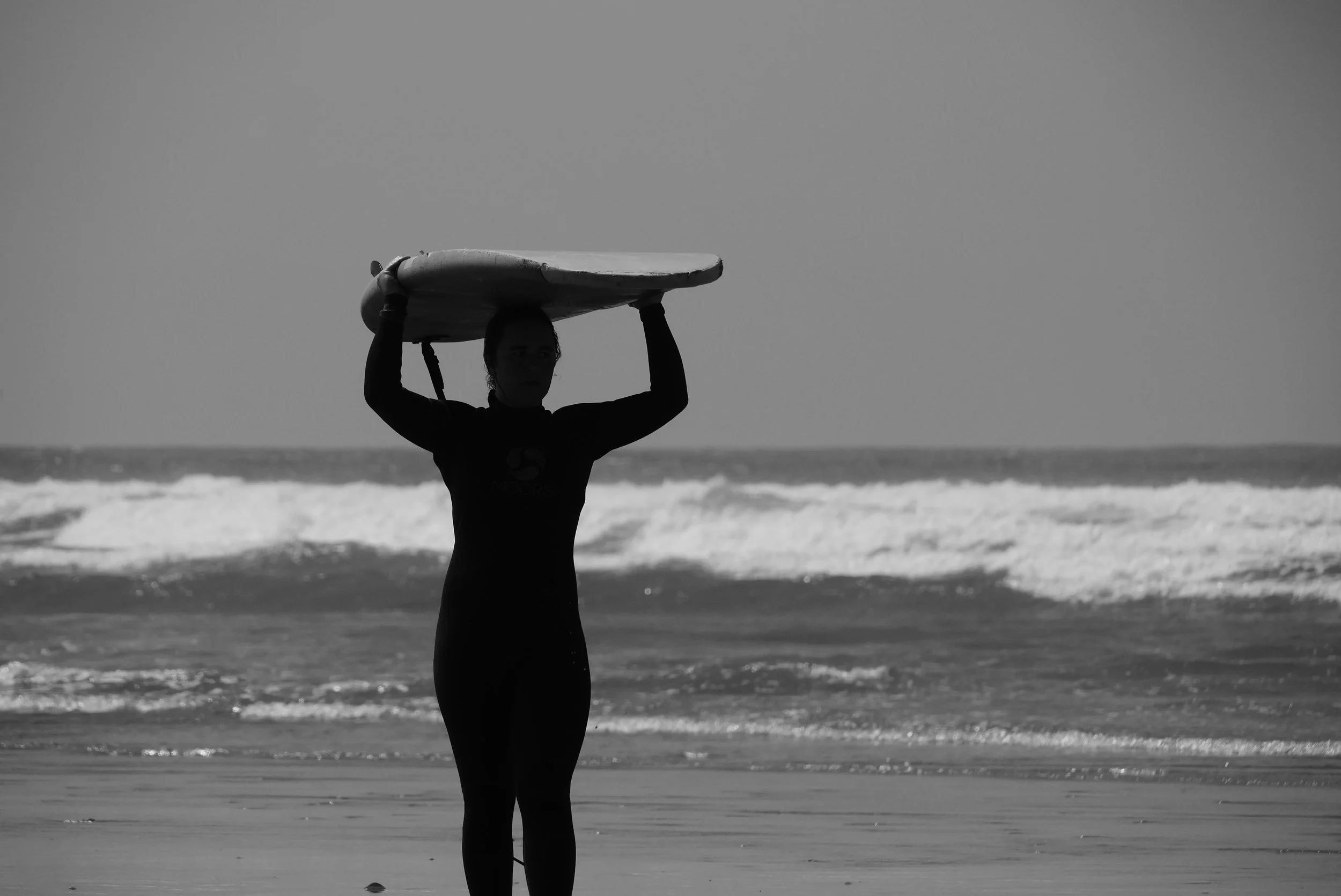 A person holding a surfboard above their head on the beach, with ocean waves in the background in black and white.