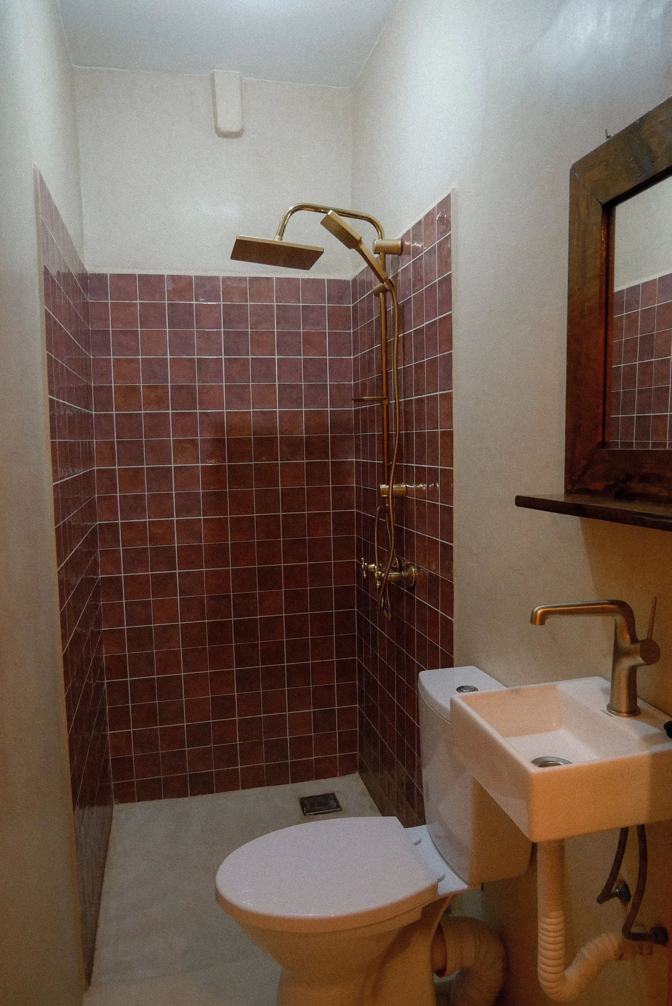 A small bathroom with a toilet, a sink, a mirror, a window, and a shower area with reddish-brown tiles and a gold-colored showerhead.
