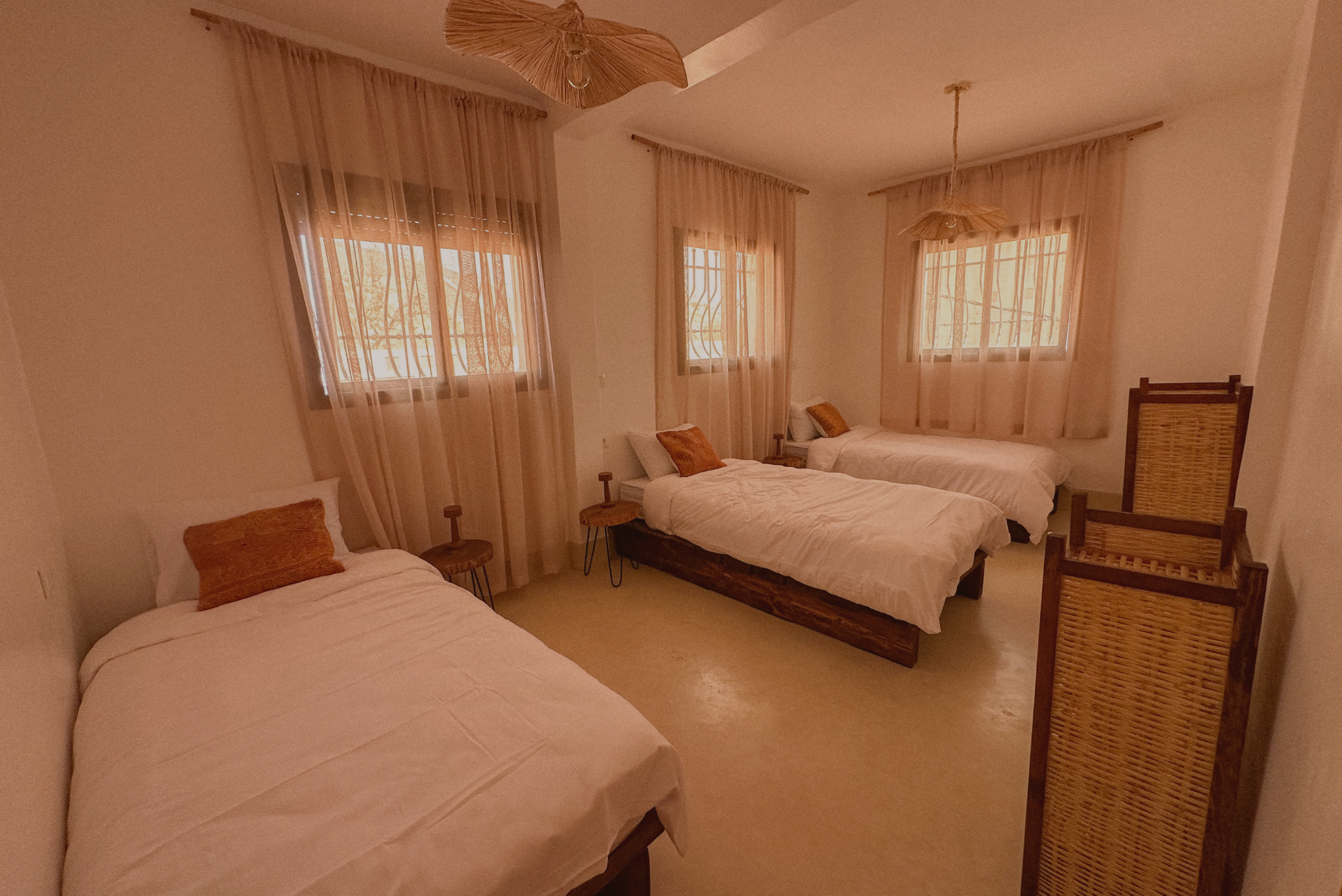 A bedroom with three beds, each with white bedding and an orange pillow. There are four windows with sheer curtains letting in natural light. The room has warm neutral colors and decorative pendant lights hanging from the ceiling.