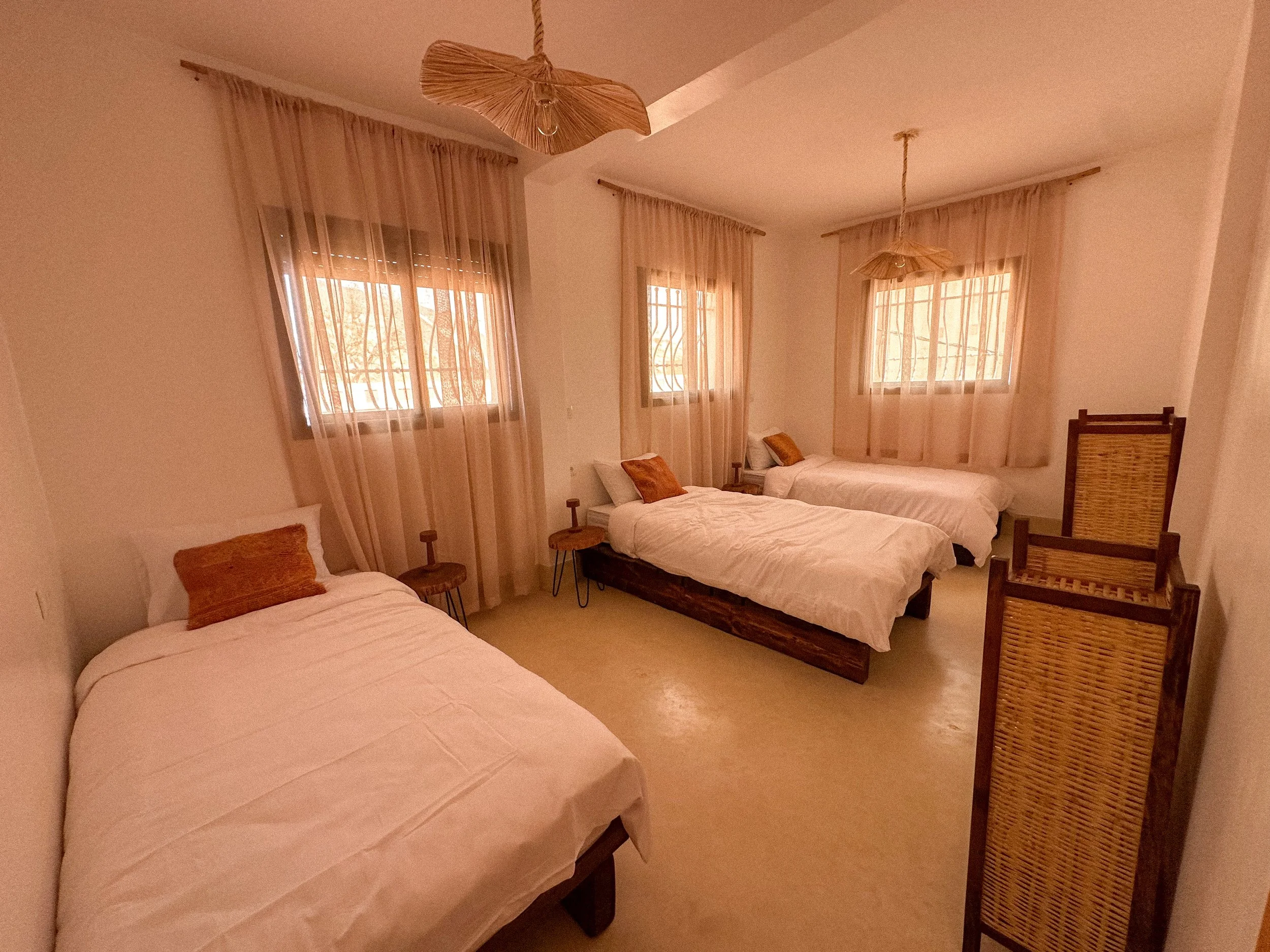 A bedroom with four single beds, each covered with a white blanket and a brown pillow. The room has three windows with sheer beige curtains, and two bamboo pendant lights hanging from the ceiling.