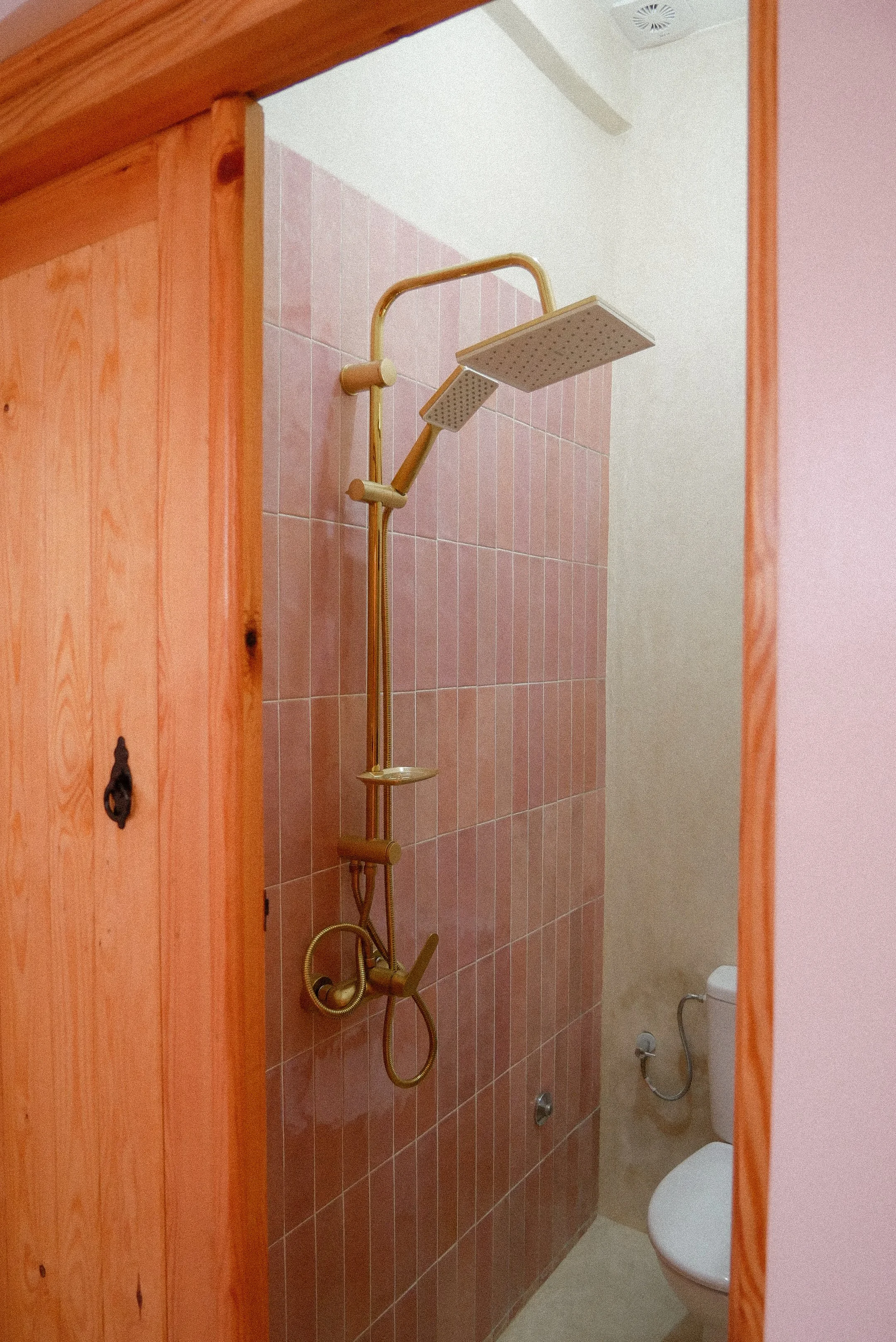 Bathroom with a brown tiled shower area, showerhead, and a toilet. The shower has a gold-colored shower pipe and fixtures. The wall separating the shower is frosted.