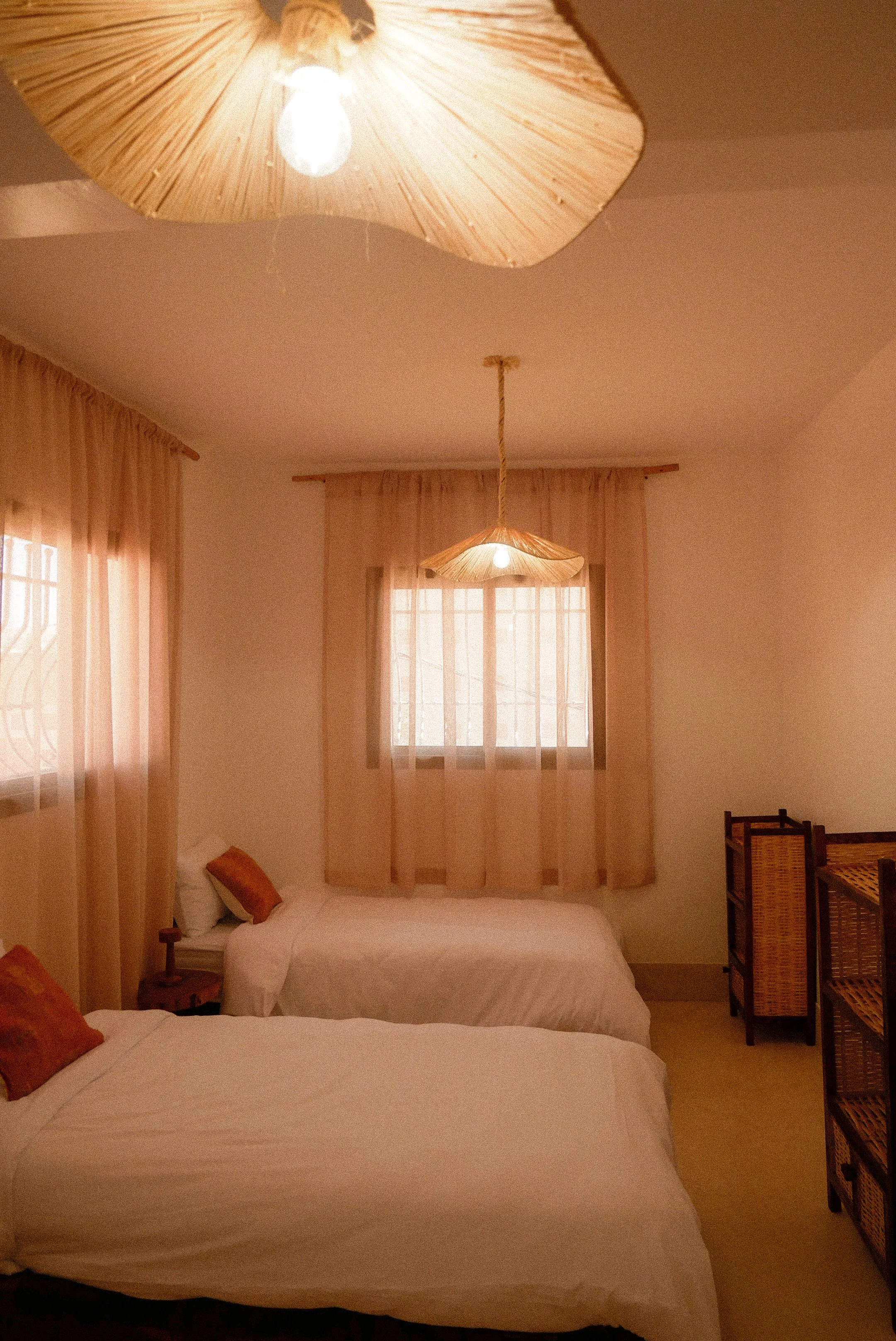 Two beds with white bedding and orange pillows in a bedroom with beige curtains and wicker furniture, ceiling light with wooden shade.