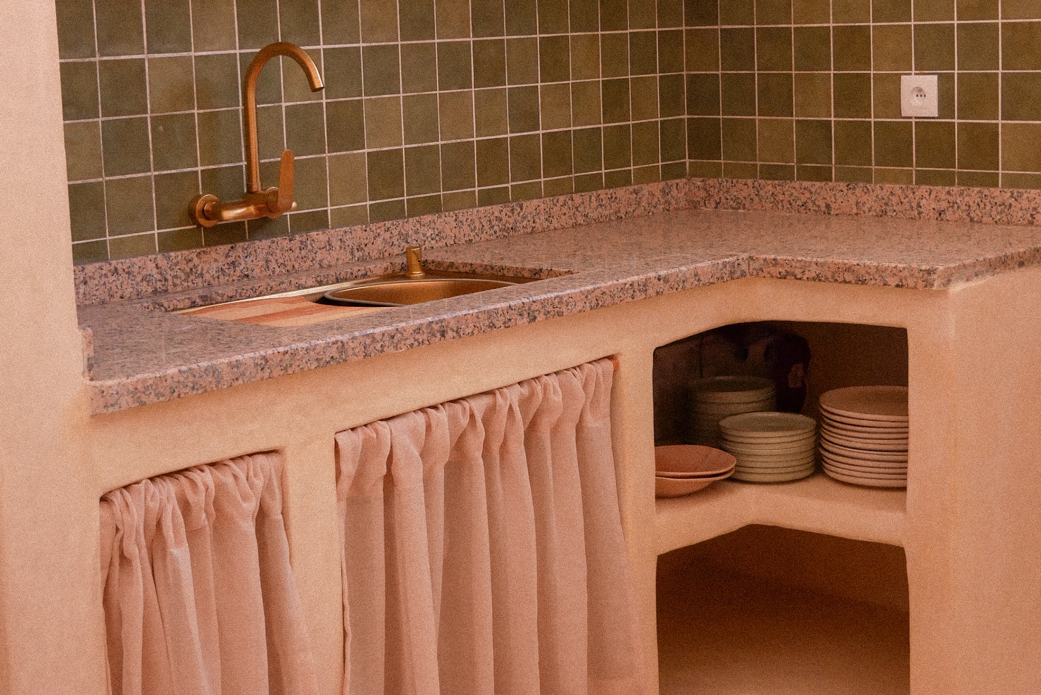 A kitchen counter with a pink and gray granite surface, green tiled backsplash, and a copper-colored faucet. Under the counter, there is a cabinet with pink ruffled curtains and a small opening revealing stacked dish plates and cups. To the right, a wall socket is visible.