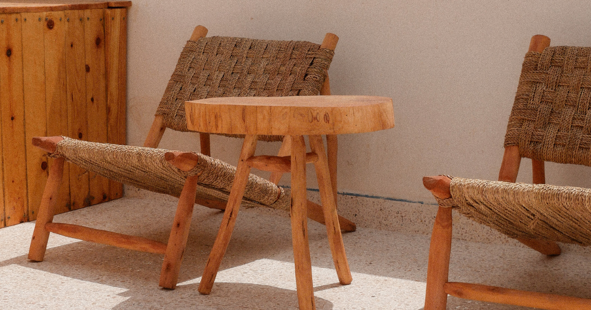 Two wooden chairs with woven seats and a small round wooden table between them, in a room with beige carpet and a wooden cabinet in the background.