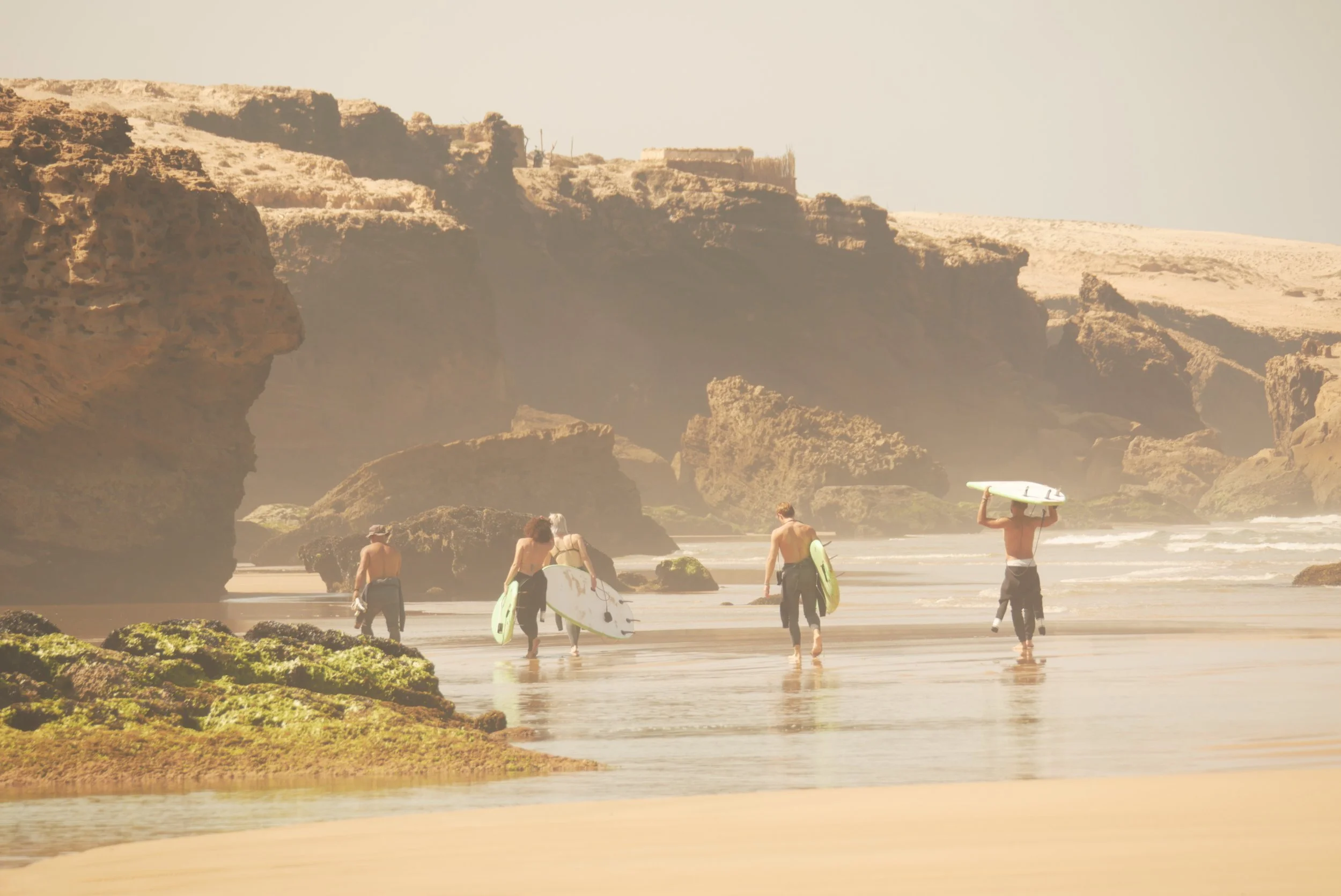 Beach scene with people carrying surfboards, large rock formations, and cliffs in the background.