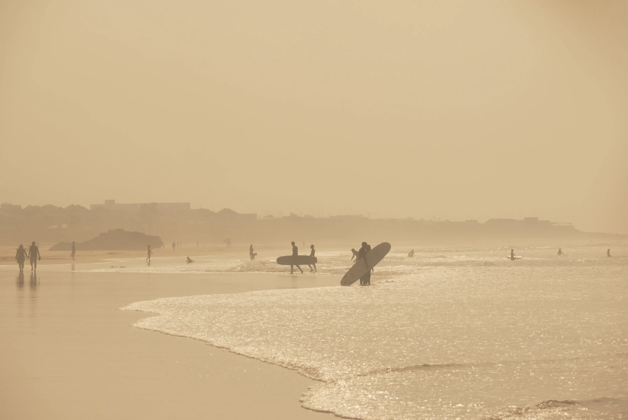 People at the beach during sunset, some with surfboards, others walking or playing in the water, with a hazy sky and distant buildings in the background.