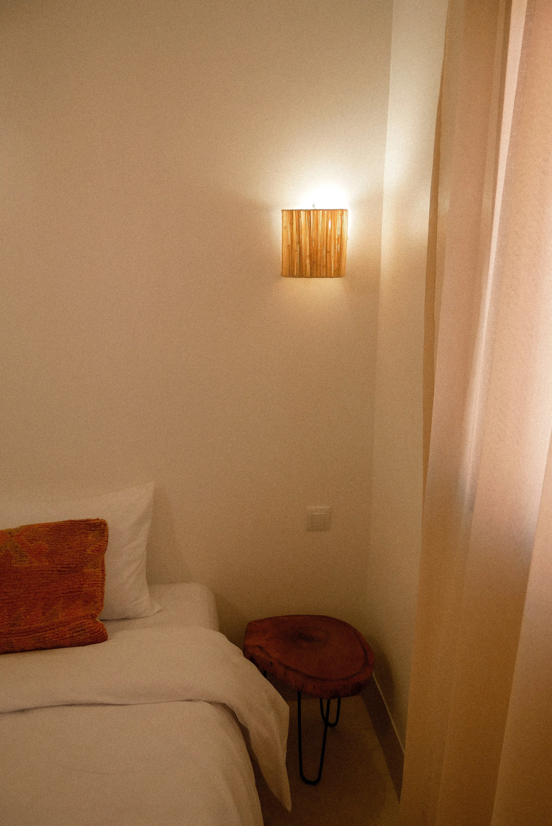 A bedroom corner with a bed that has white sheets, a brown textured pillow, and a second plain white pillow. Next to the bed is a small round wooden side table with angled black metal legs. A beige wall with a wooden wall lamp is visible, casting light on the wall.
